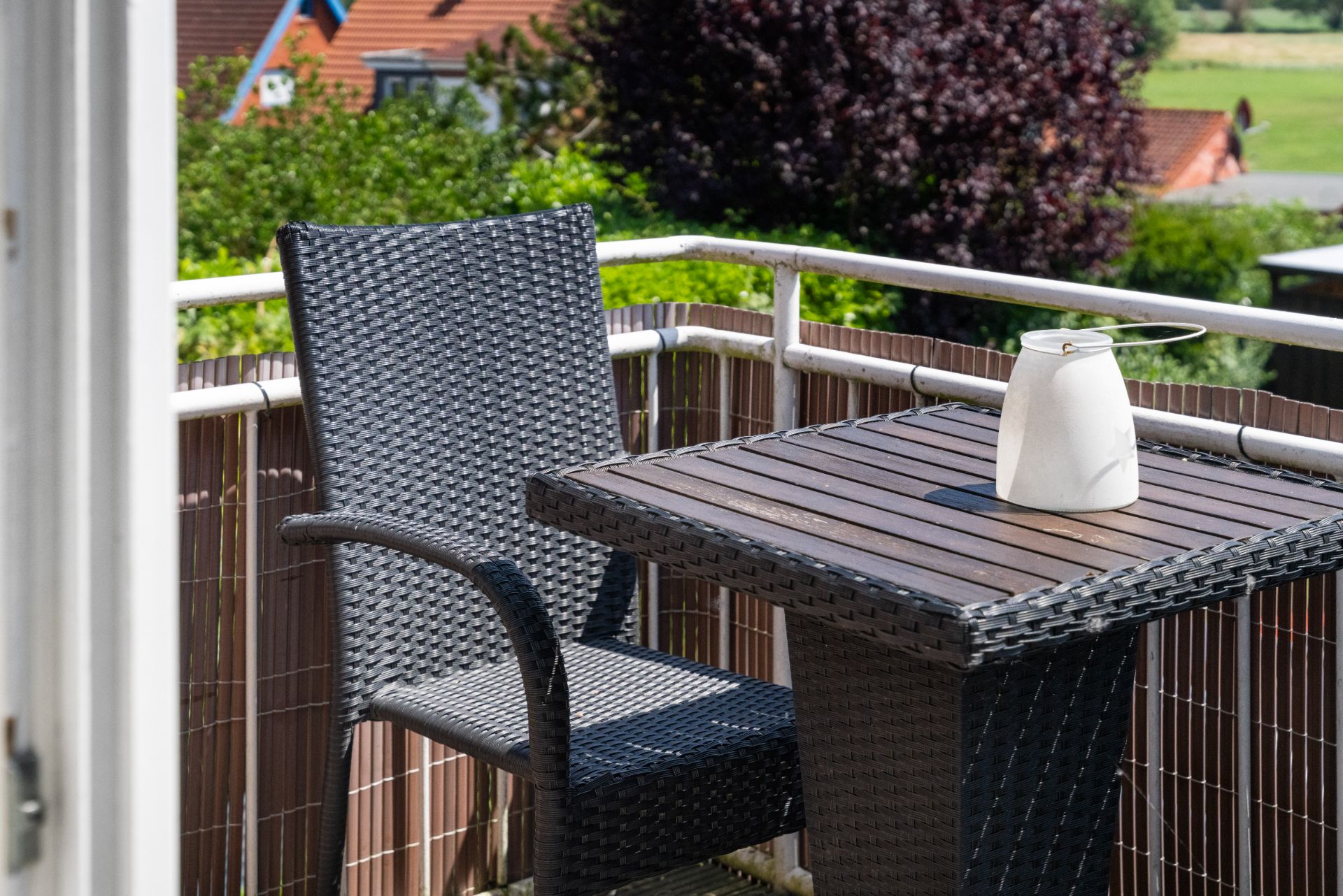 Balkon mit Rattanstuhl, Tisch und weißer Vase, im Hintergrund Garten und Hausdächer.