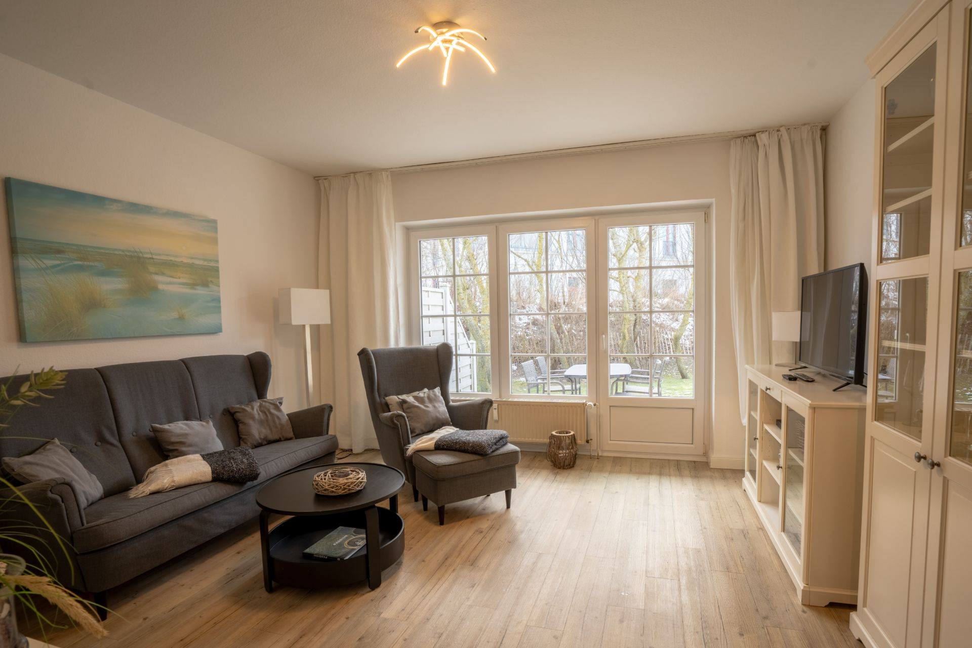 Helles Wohnzimmer mit Sofa, Sessel, TV, Couchtisch und großem Fenster zum Garten.