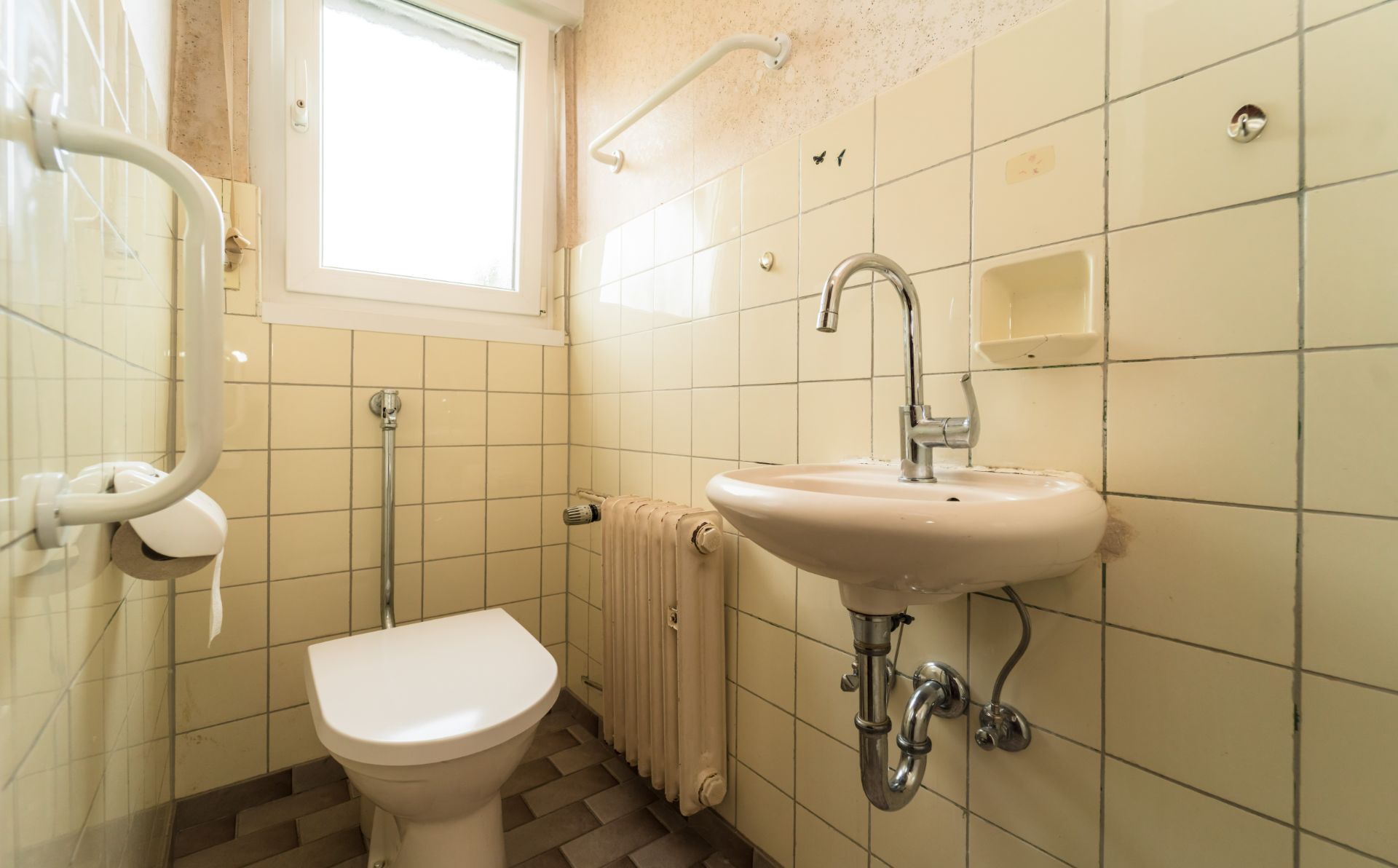 Kleines Bad mit Toilette, Waschbecken, Heizung und Haltegriffen, gelbe Fliesen und Fenster.