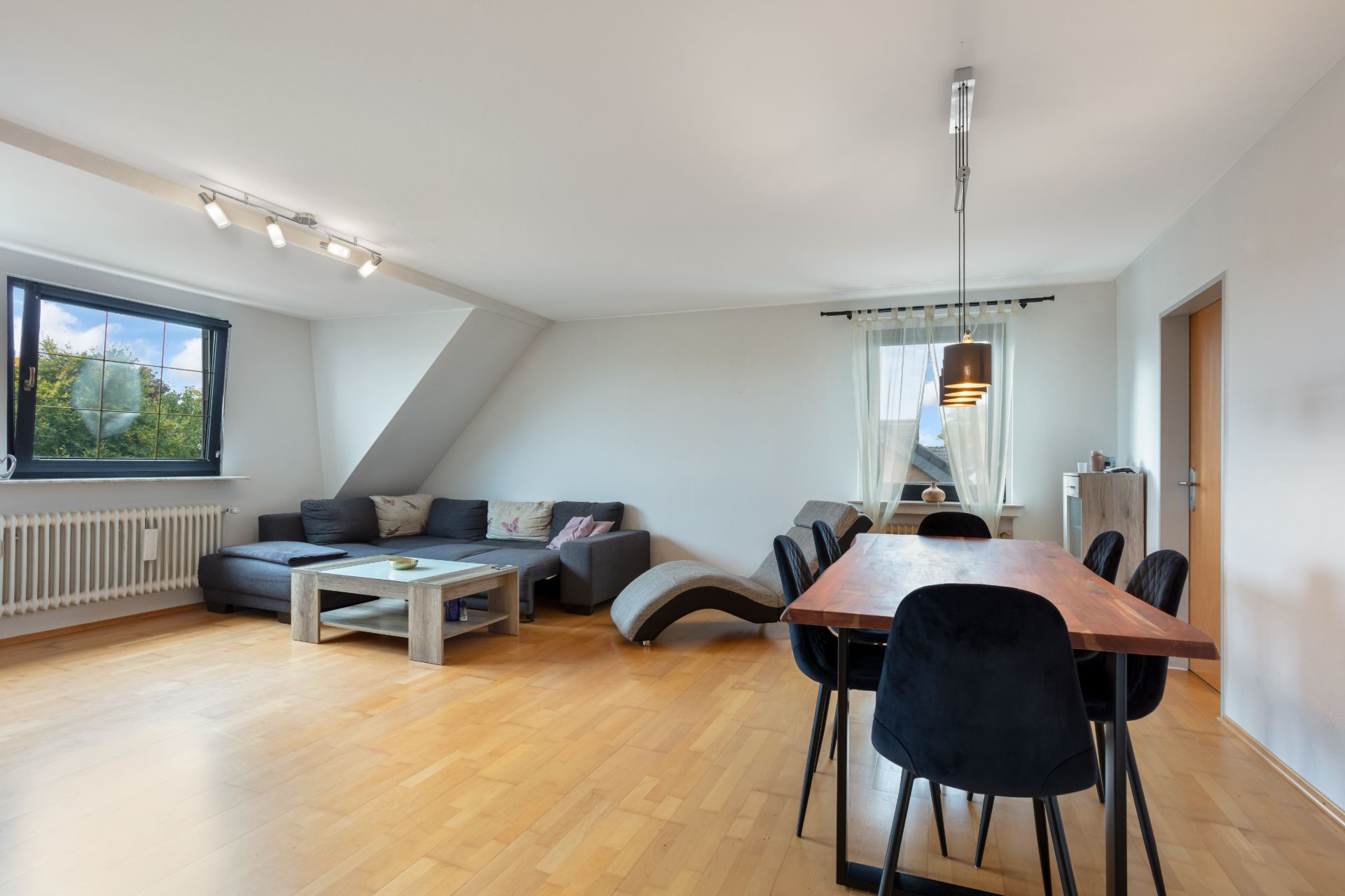 Wohnzimmer mit Ecksofa, Holztisch, Essbereich und großen Fenstern auf hellem Holzboden.