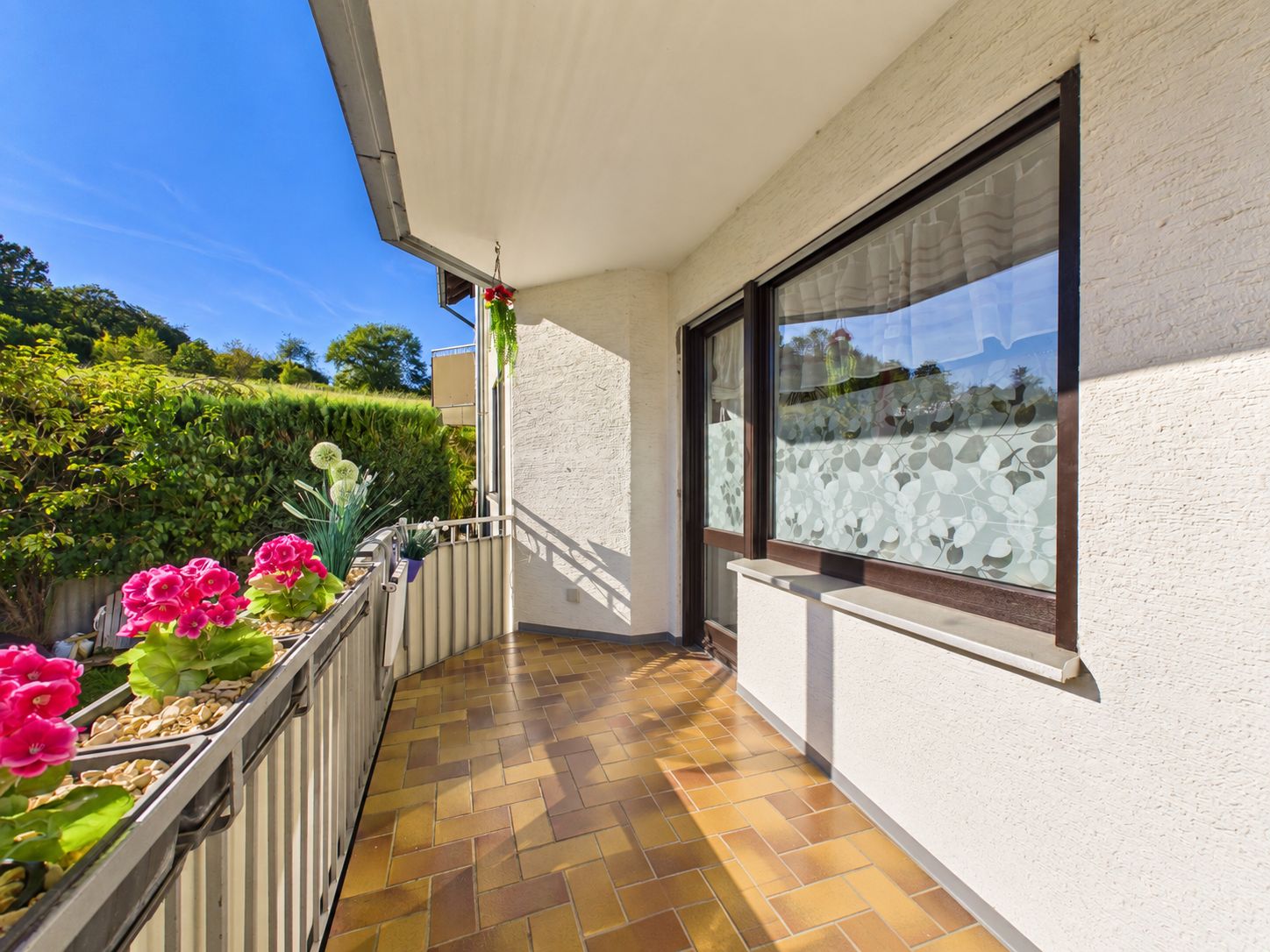 Überdachter Balkon mit Blumen auf dem Geländer und großen Fenstern zur Wohnung.