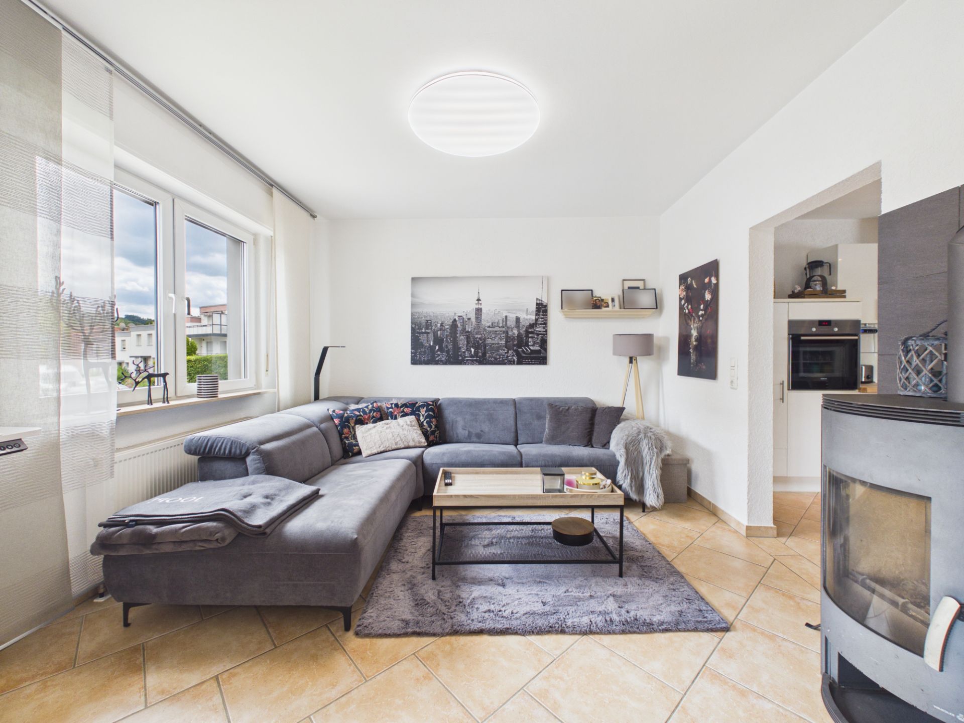 Helles Wohnzimmer mit grauem Ecksofa, Couchtisch, Kaminofen und großem Fenster.