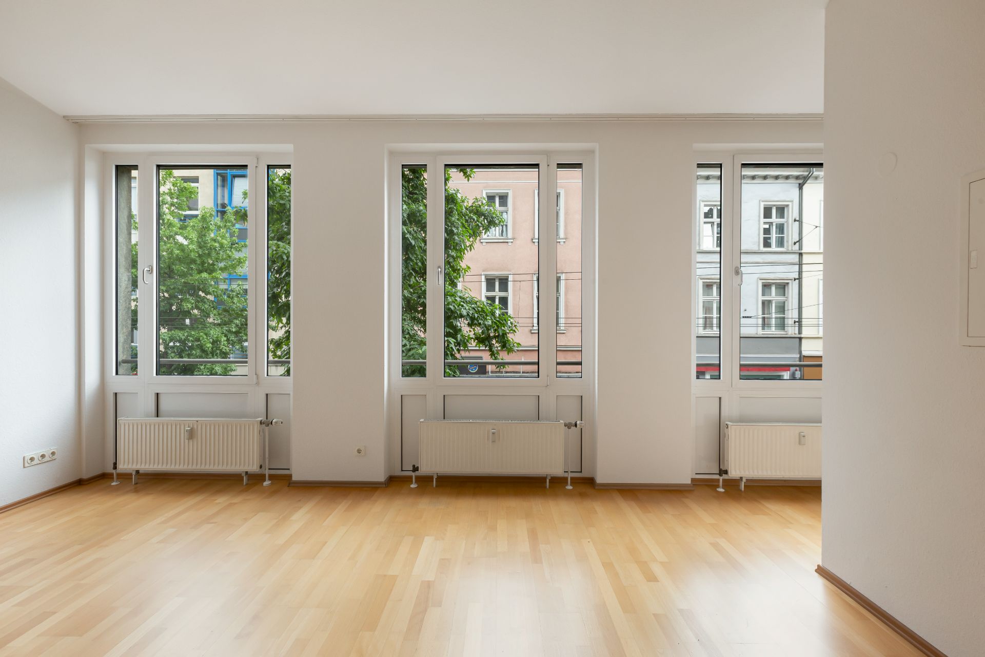 Helles, leeres Zimmer mit Parkettboden und großen Fenstern zur Straße.