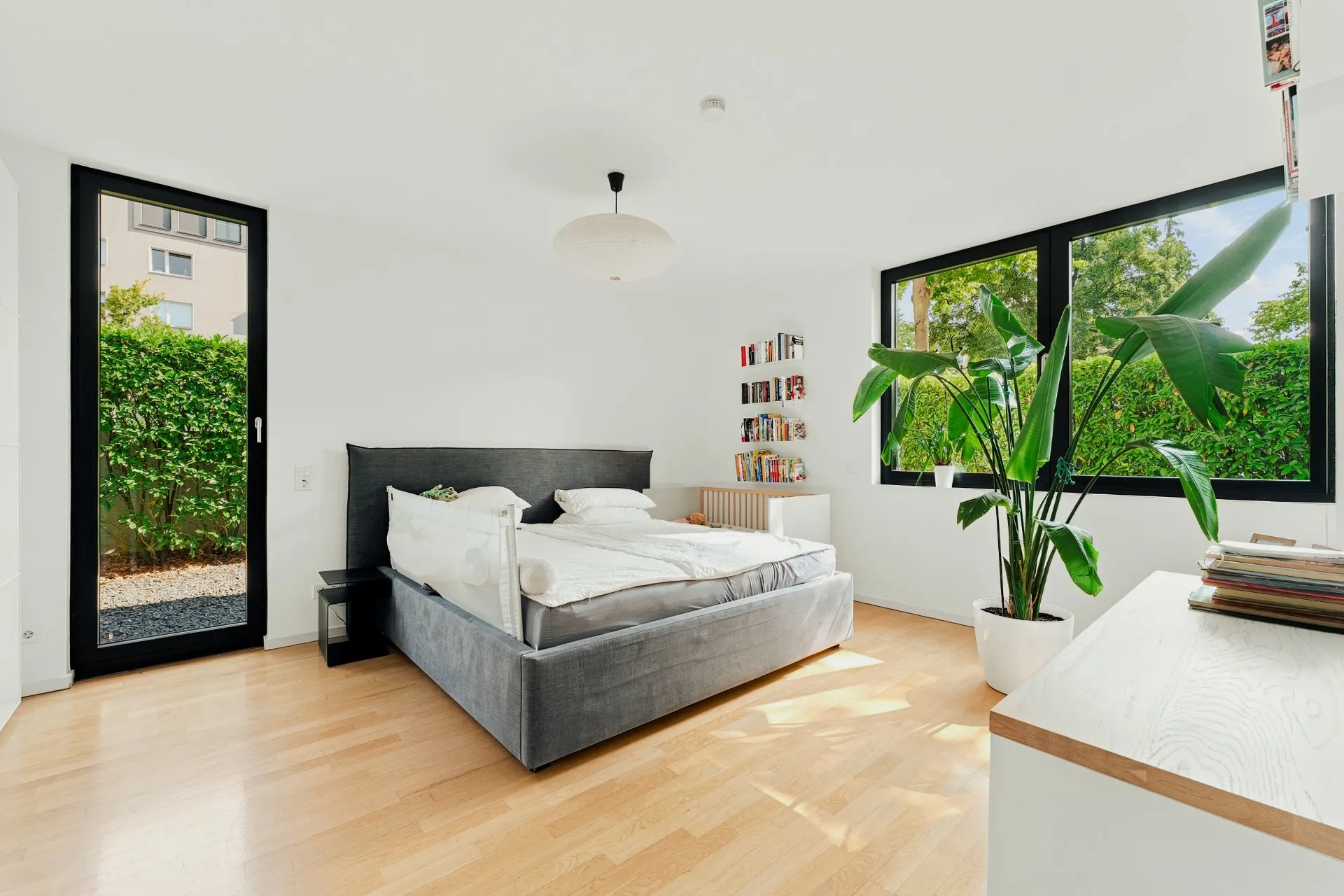 Modernes Schlafzimmer mit grauem Bett, großen Fenstern, Holzboden und großer Zimmerpflanze.