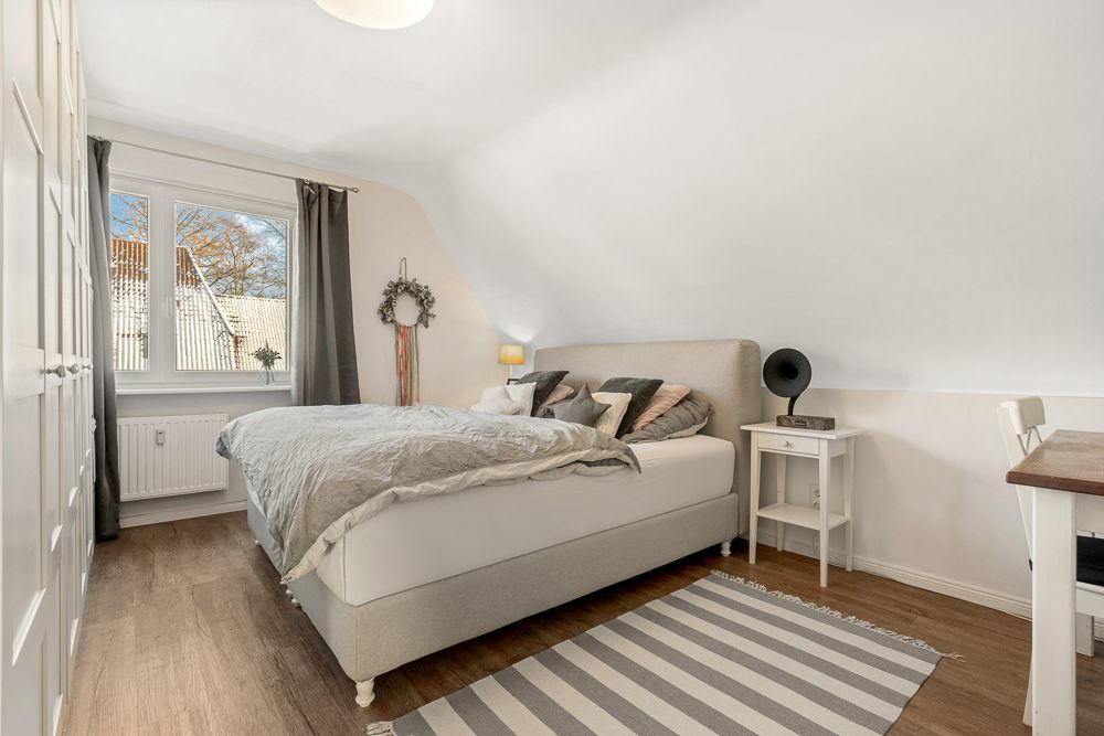 Helles Schlafzimmer mit Doppelbett, Schreibtisch, Teppich und großem Fenster mit Vorhängen.