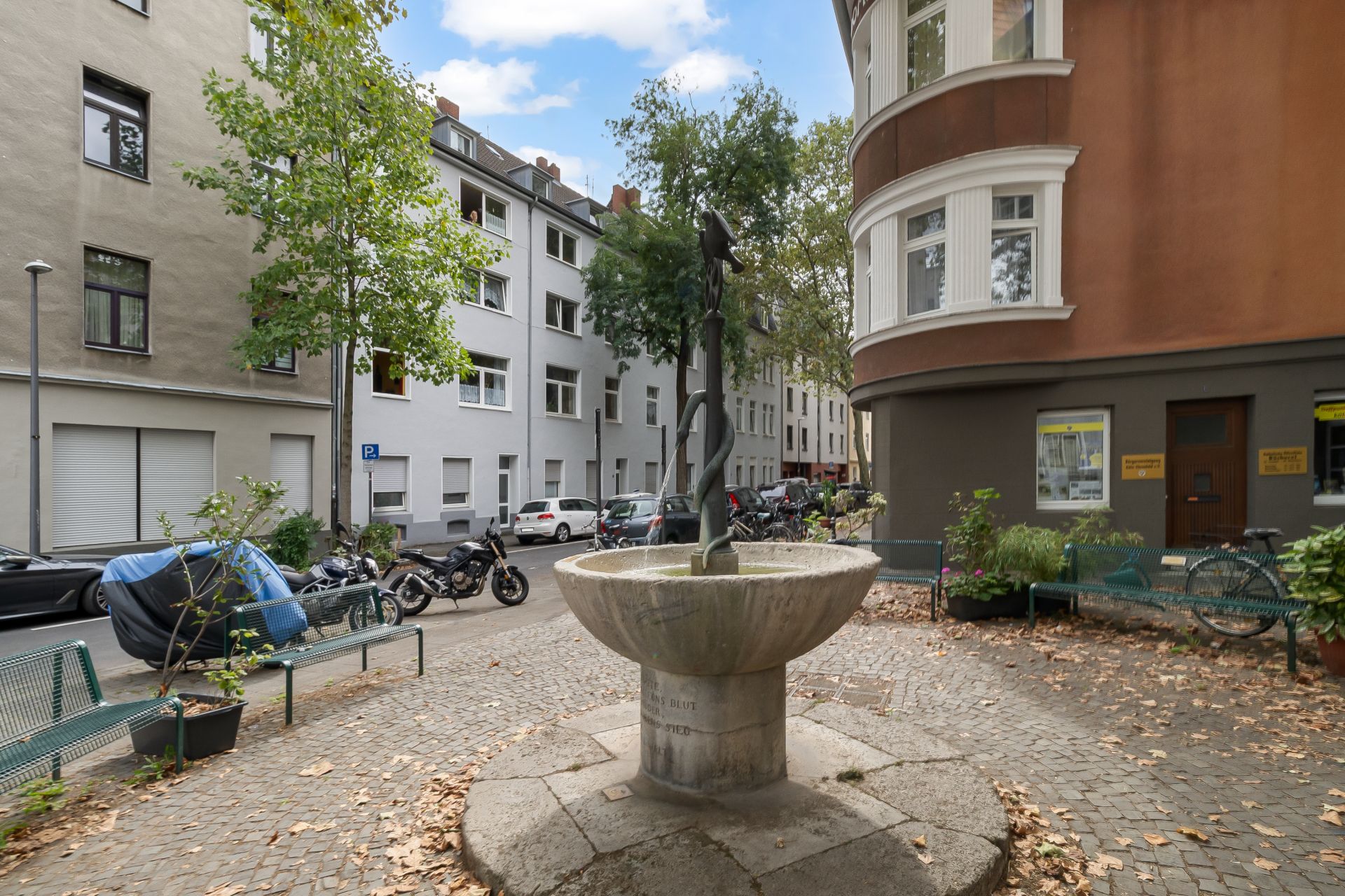 Kleiner Platz mit Brunnen, Bänken und Bäumen zwischen Wohnhäusern an einer ruhigen Straße.