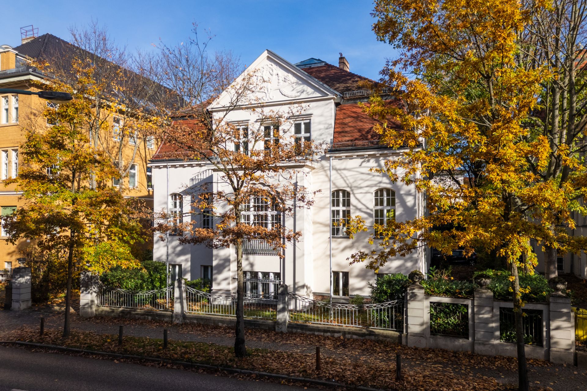 Weißes Wohnhaus mit roten Dachziegeln, umgeben von Bäumen mit Herbstlaub.