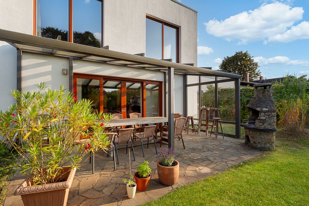 Überdachte Terrasse mit Esstisch, Stühlen, Pflanzkübeln und gemauertem Grill im Garten.