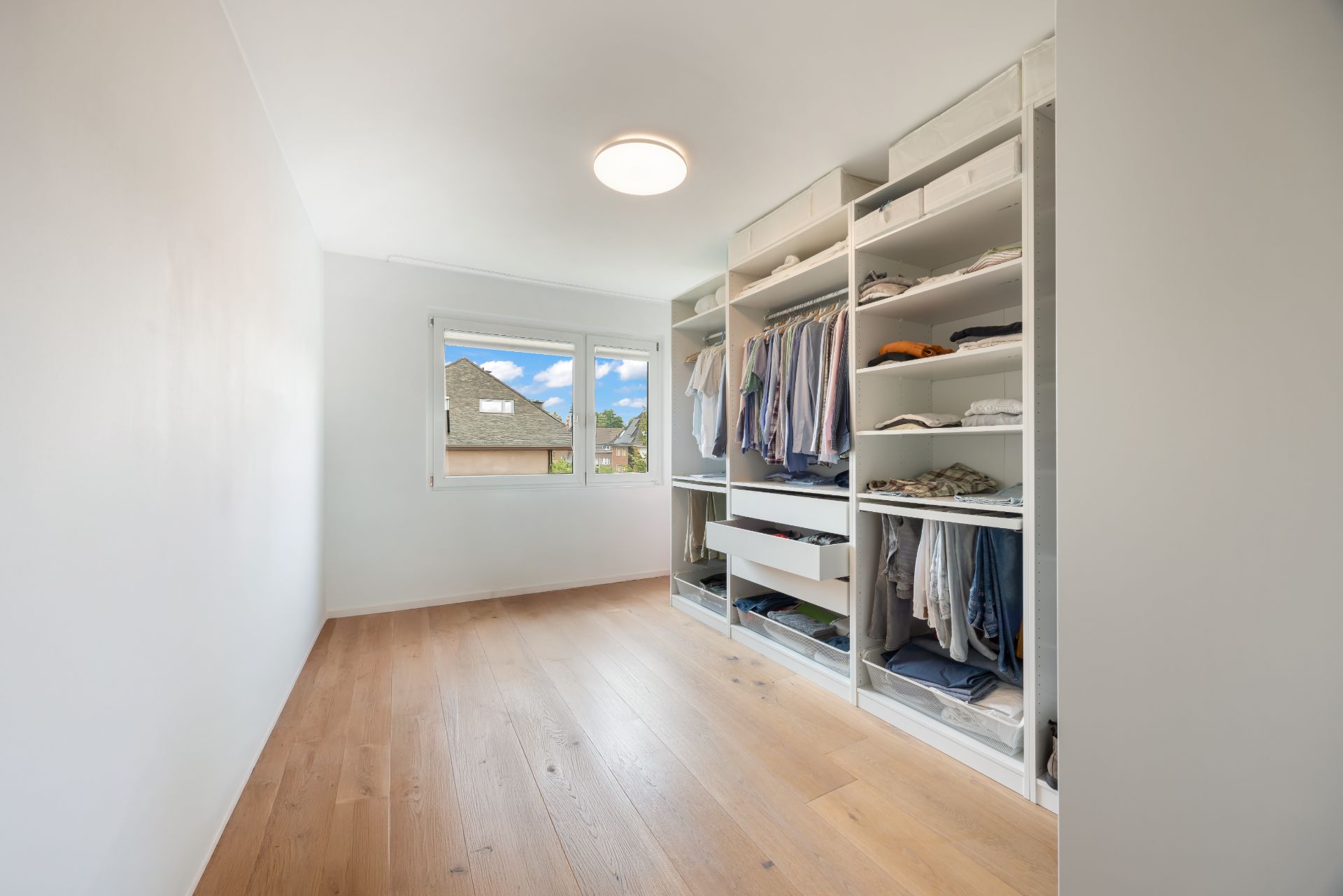 Heller Ankleideraum mit offenem Kleiderschrank, Holzfußboden und Fenster mit Ausblick.