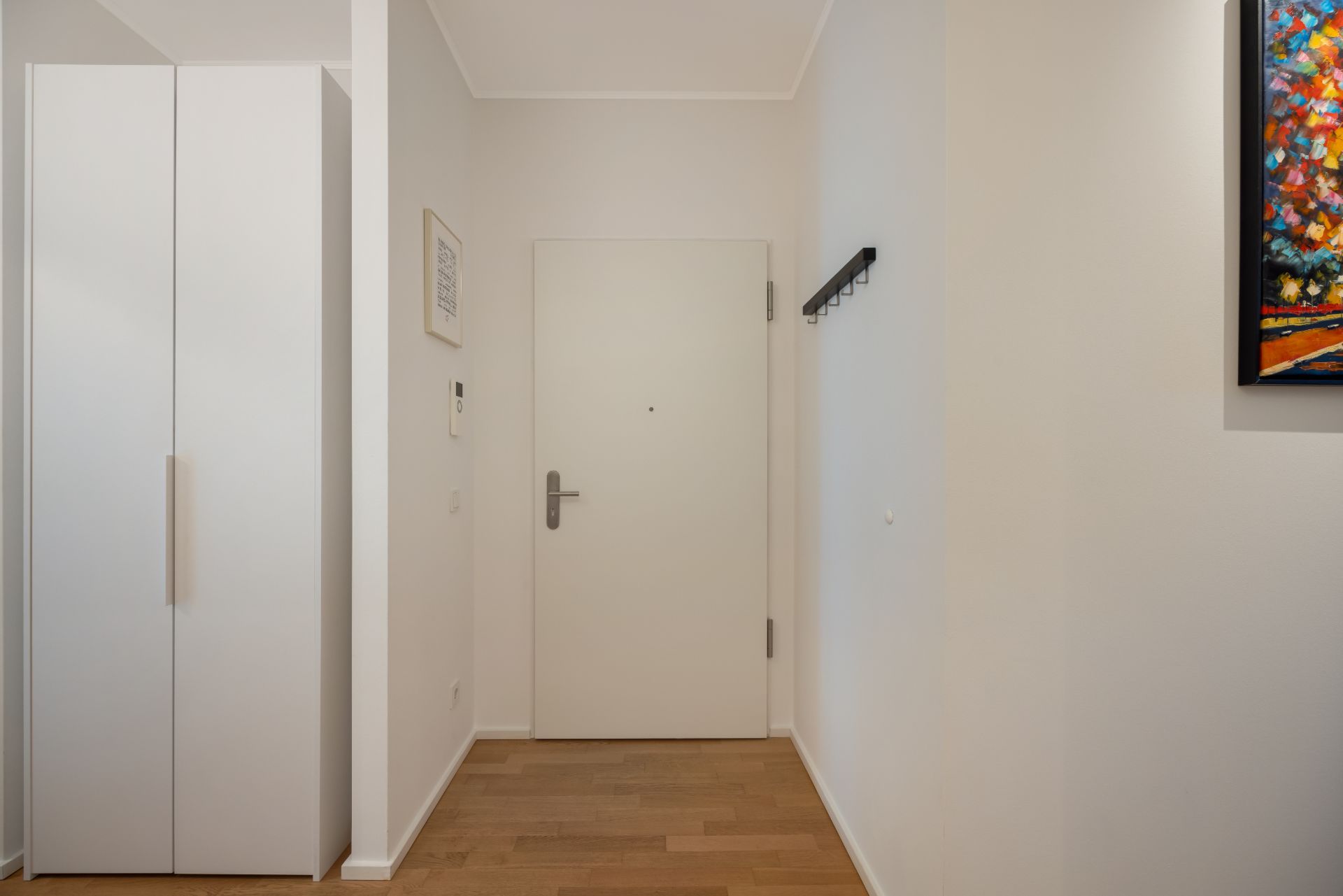 Heller Flur mit weißer Tür, weißem Schrank, Garderobenleiste und buntem Bild an der Wand.