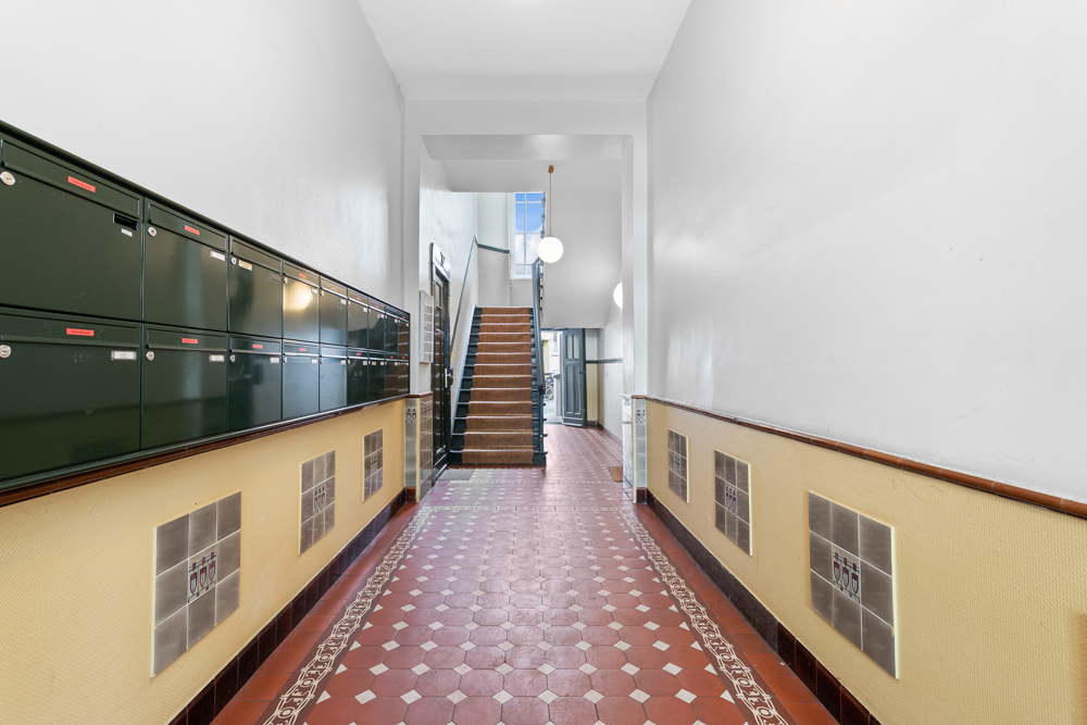 Hausflur mit roten Bodenfliesen, Briefkästen an der Wand und einer Treppe im Hintergrund.