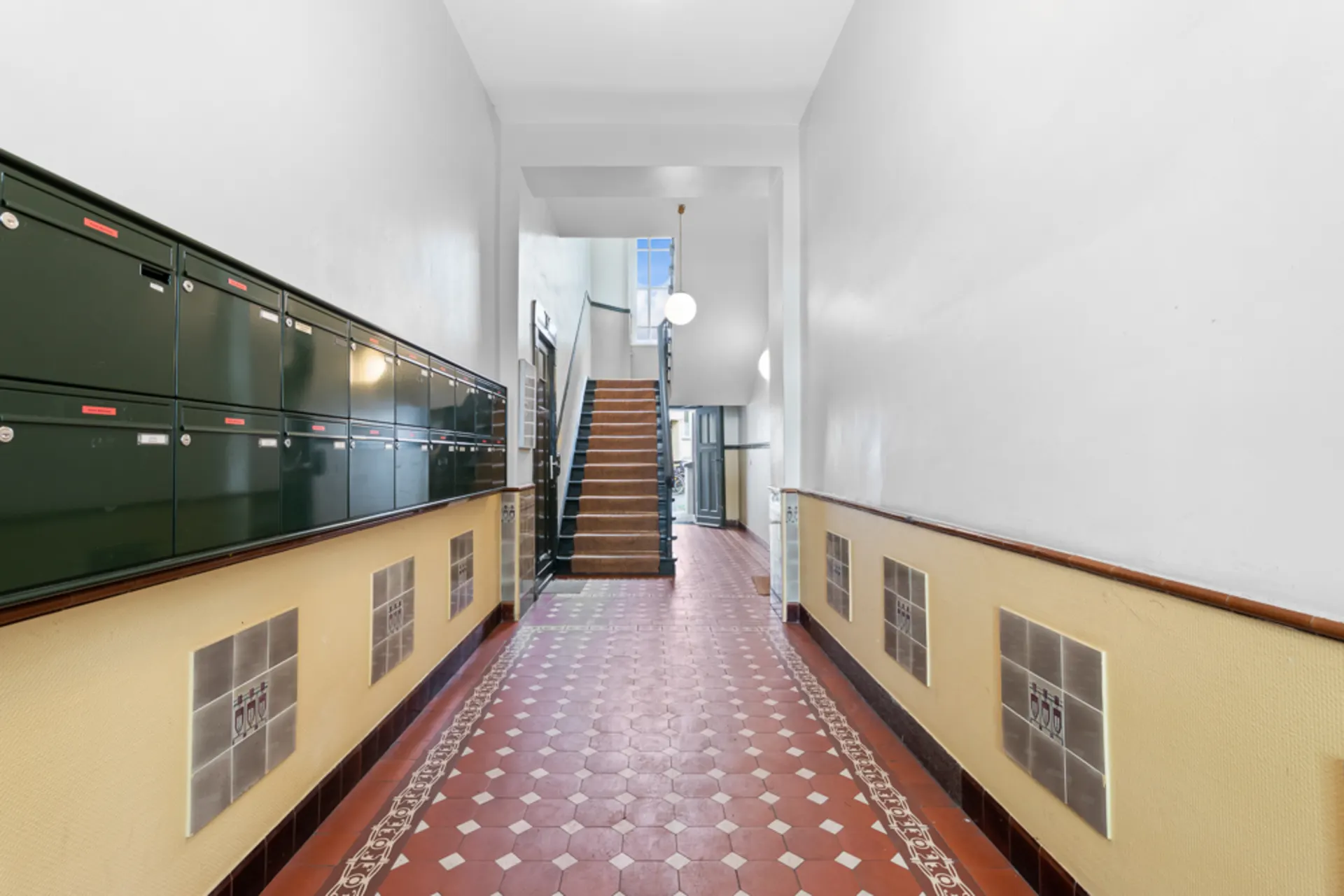 Hausflur mit roten Bodenfliesen, Briefkästen an der Wand und einer Treppe im Hintergrund.