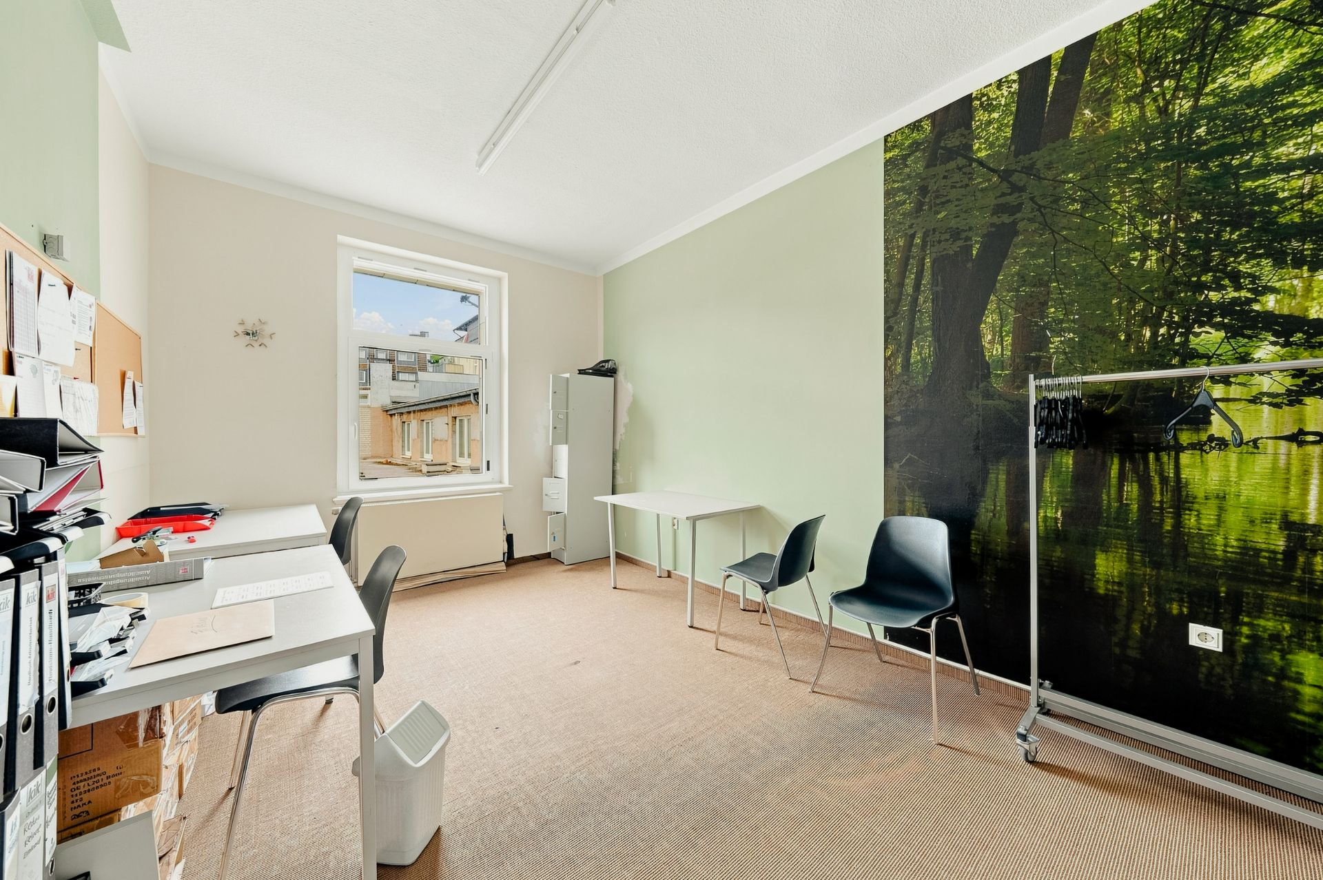 Helles Arbeitszimmer mit Schreibtischen, Stühlen, Regalen und Wandbild eines Waldes.