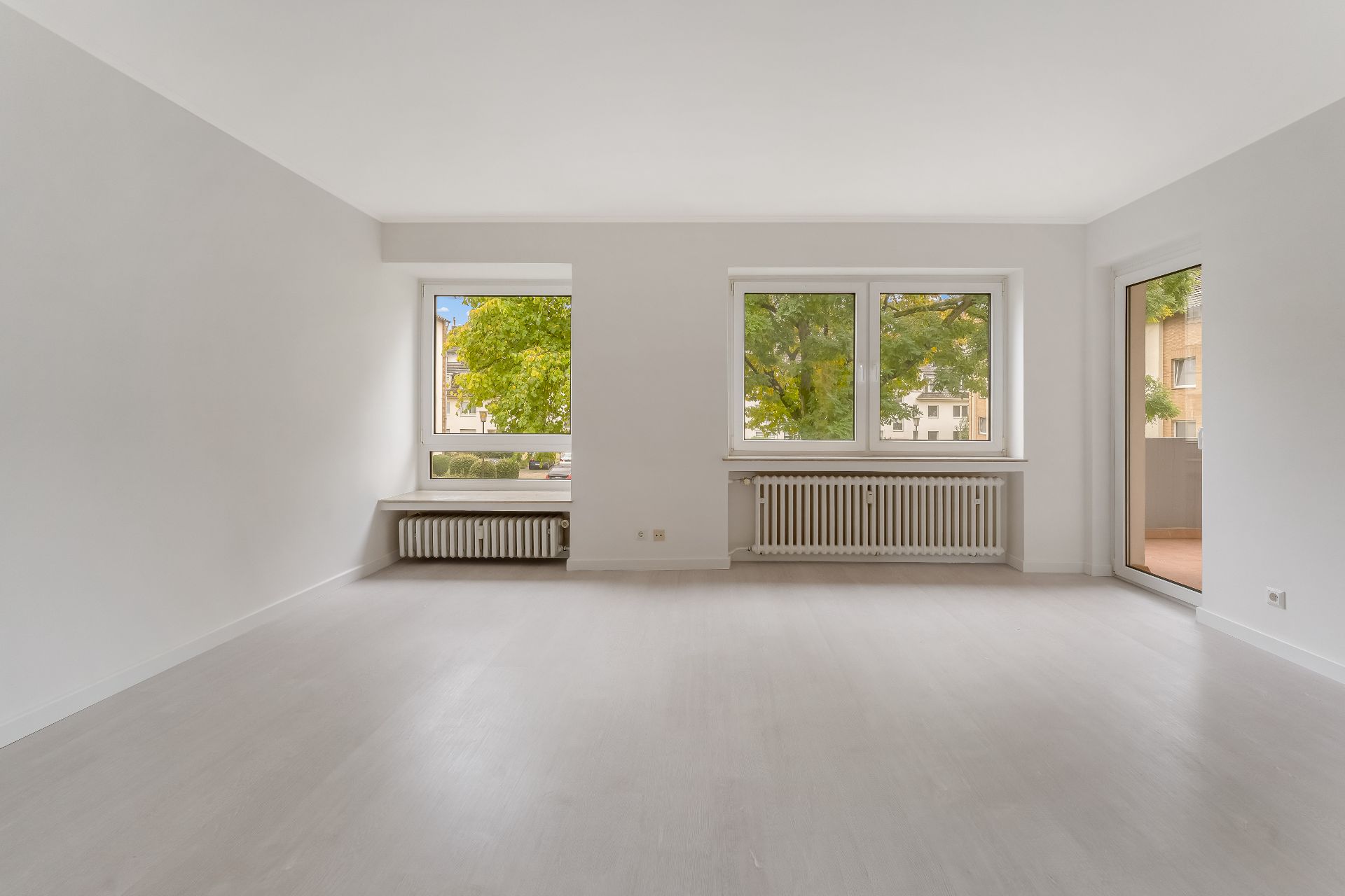 Helles, leeres Zimmer mit zwei großen Fenstern, Heizkörpern und Zugang zu einem Balkon.