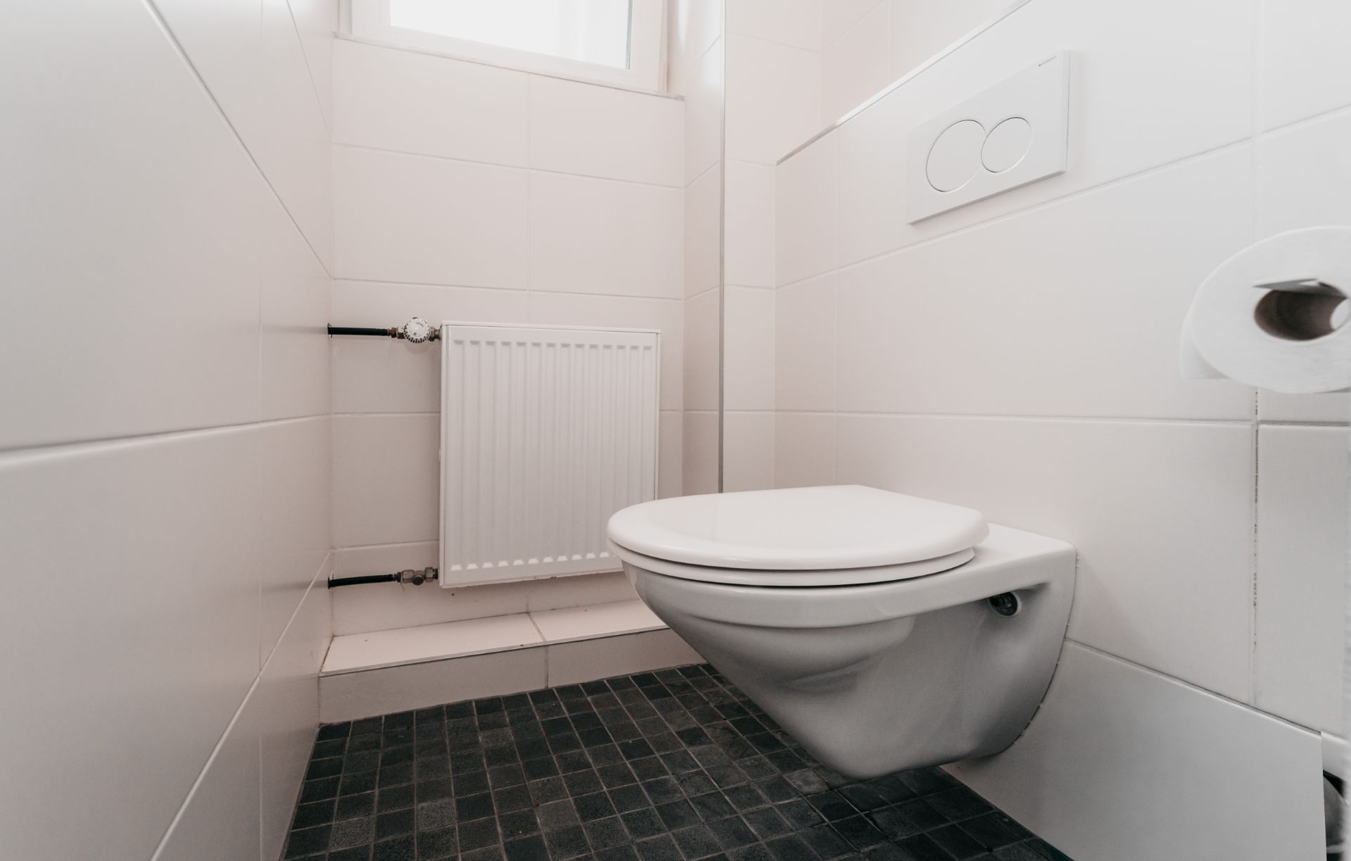 Kleines, weiß gefliestes Bad mit Wandtoilette, Heizung und Fenster.