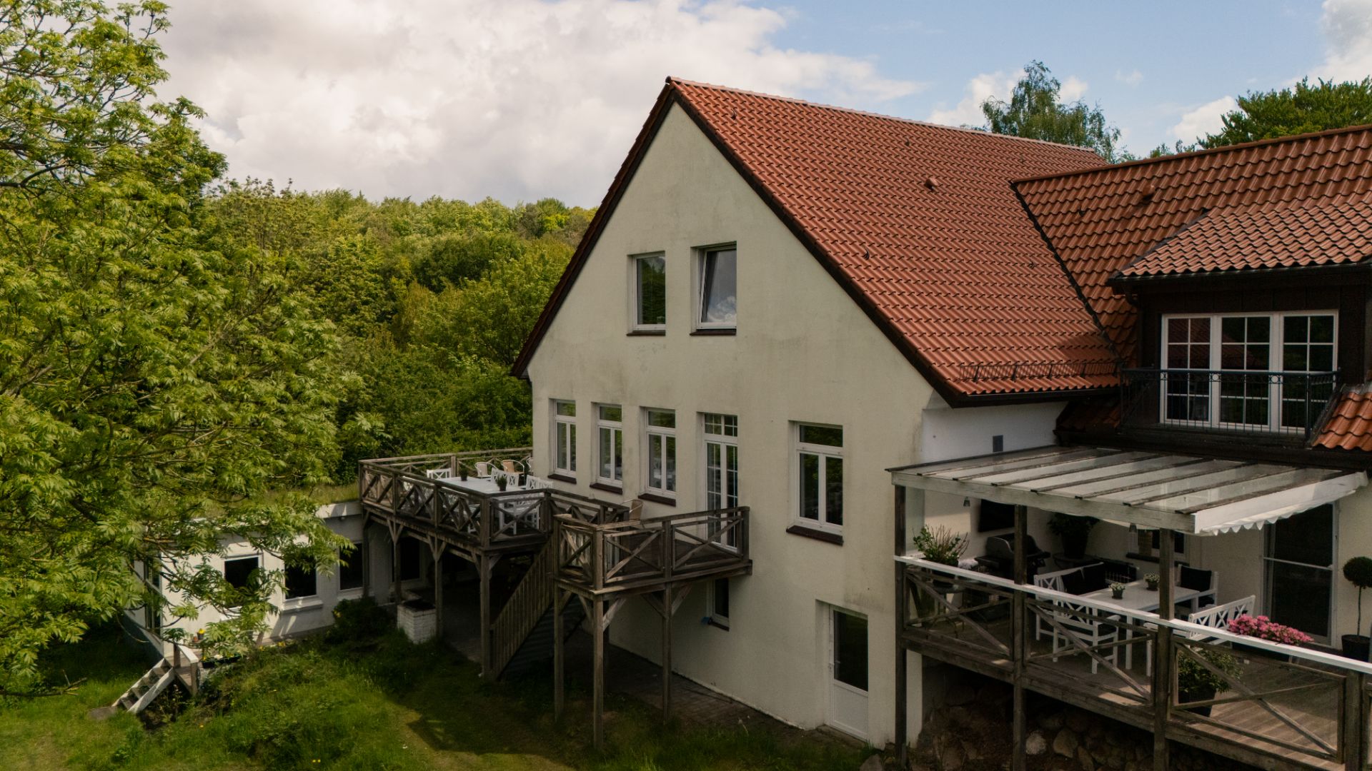 Zweistöckiges Haus mit rotem Dach, Holzterrassen und Blick auf einen Garten mit Bäumen.