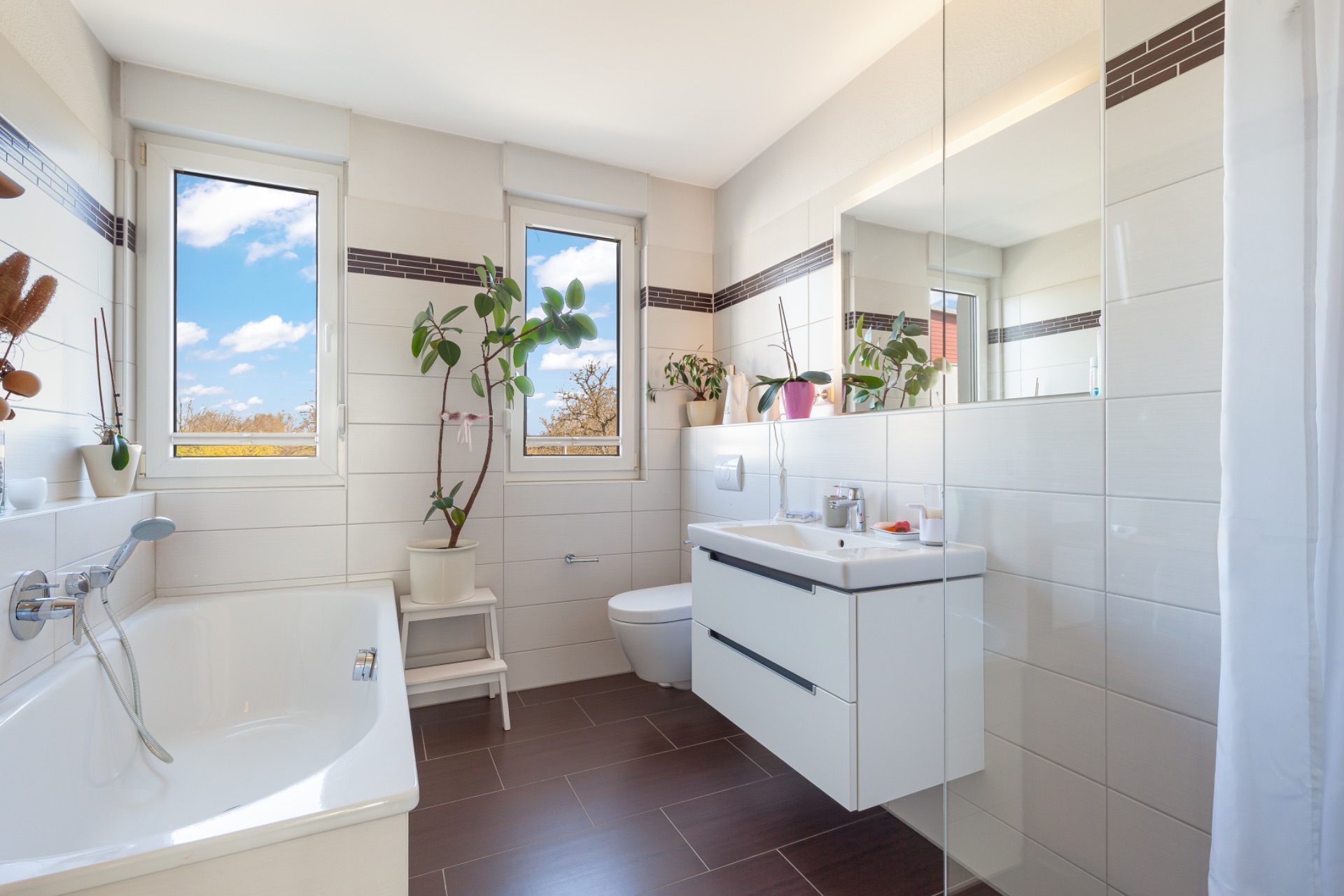 Modernes Badezimmer mit Badewanne, zwei Fenstern, WC, Waschbecken und Pflanzen.
