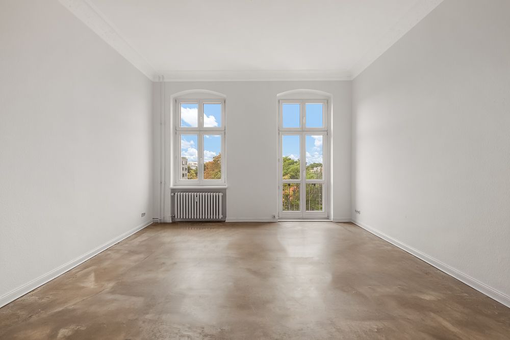 Leerer Raum mit großen Fenstern, weiß gestrichenen Wänden und braunem Boden.