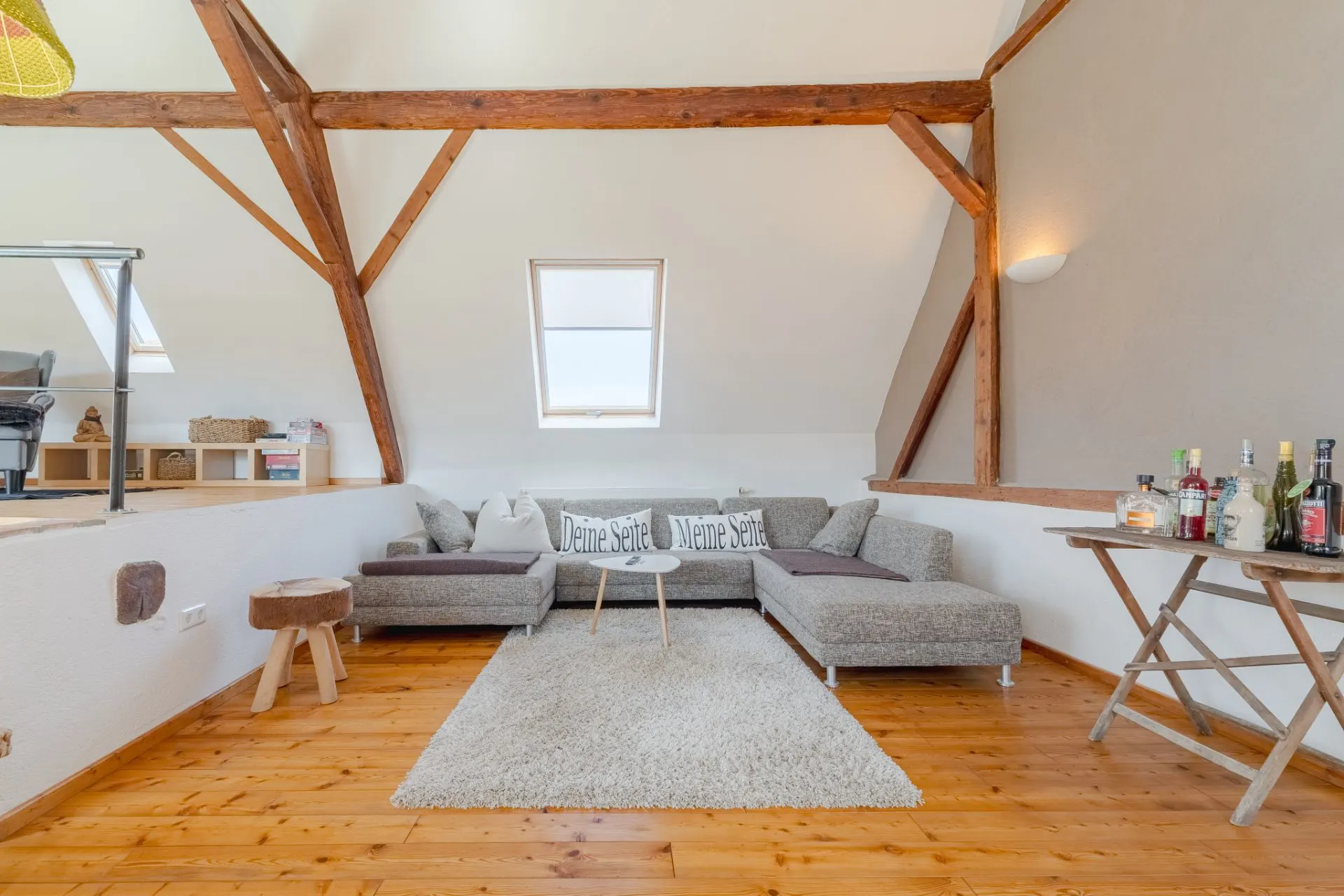 Wohnzimmer mit grauem Ecksofa, Teppich, Holzboden und sichtbaren Dachbalken.