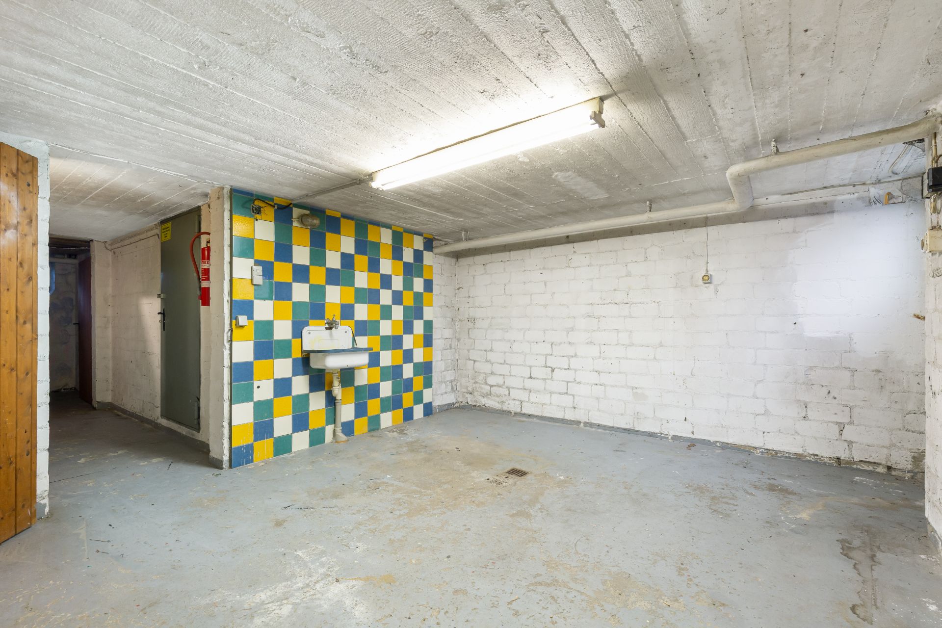 Kellerraum mit bunter Fliesenwand und Waschbecken, weiße Wände und Betonboden.