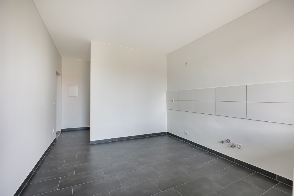 Leerer, heller Raum mit grauem Fliesenboden und gefliester Wand, Anschlüsse sichtbar.