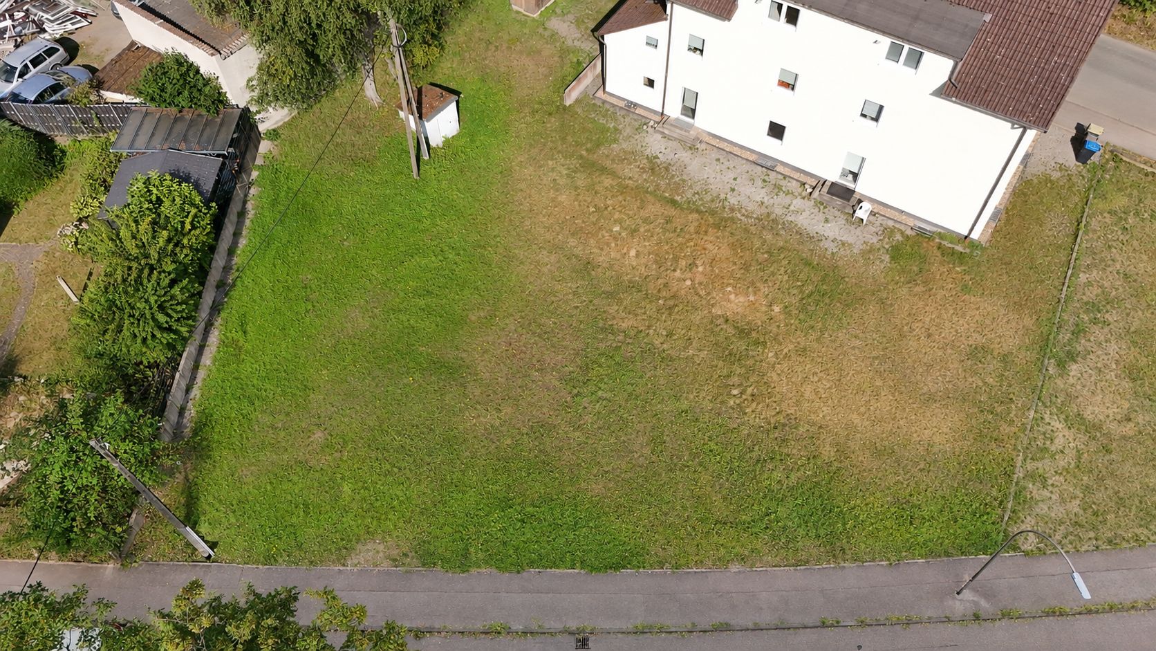 Luftaufnahme eines grünen Gartengrundstücks neben einem weißen Wohnhaus an einer Straße.