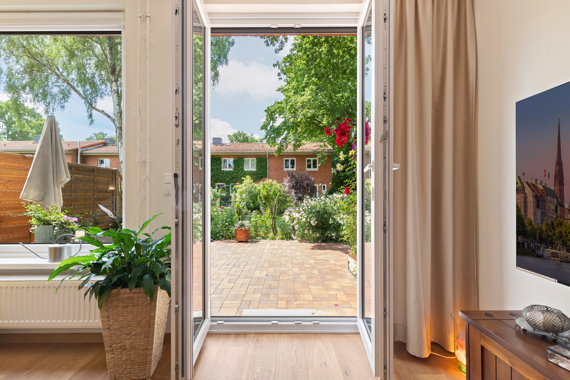 Blick durch geöffnete Terrassentür auf einen Garten mit Blumen und Ziegelterrasse.