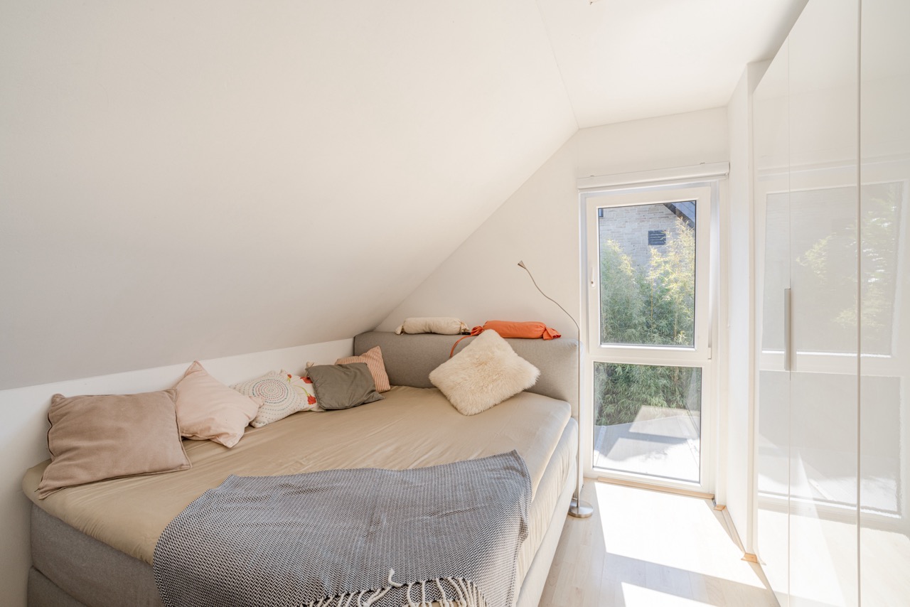 Helles Zimmer mit Bett, vielen Kissen, großem Fenster und weißem Schrank unter einer Dachschräge.