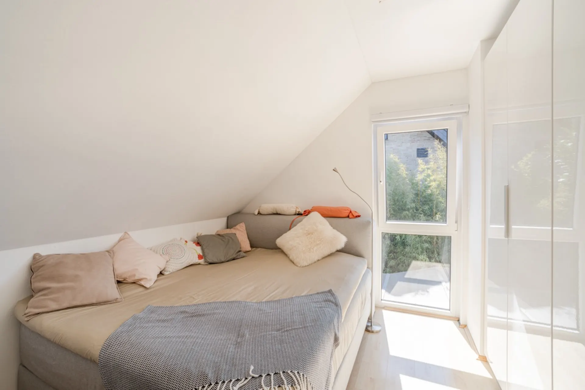 Helles Zimmer mit Bett, vielen Kissen, großem Fenster und weißem Schrank unter einer Dachschräge.