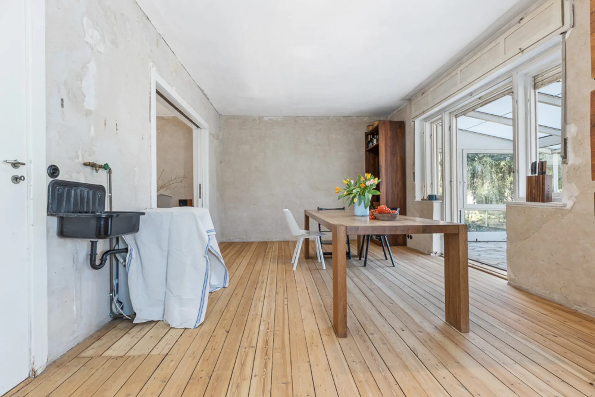 Heller Raum mit Holzboden, Esstisch, Stühlen, Blumen und großem Fenster, Wand mit Waschbecken.