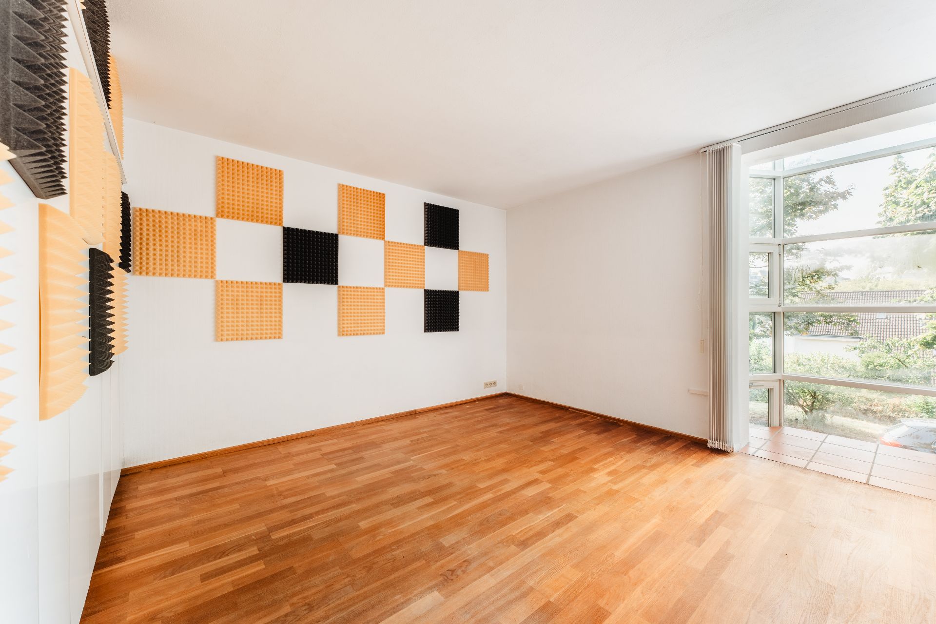 Leerer Raum mit Holzfußboden, großen Fenstern und Akustikplatten an der weißen Wand.