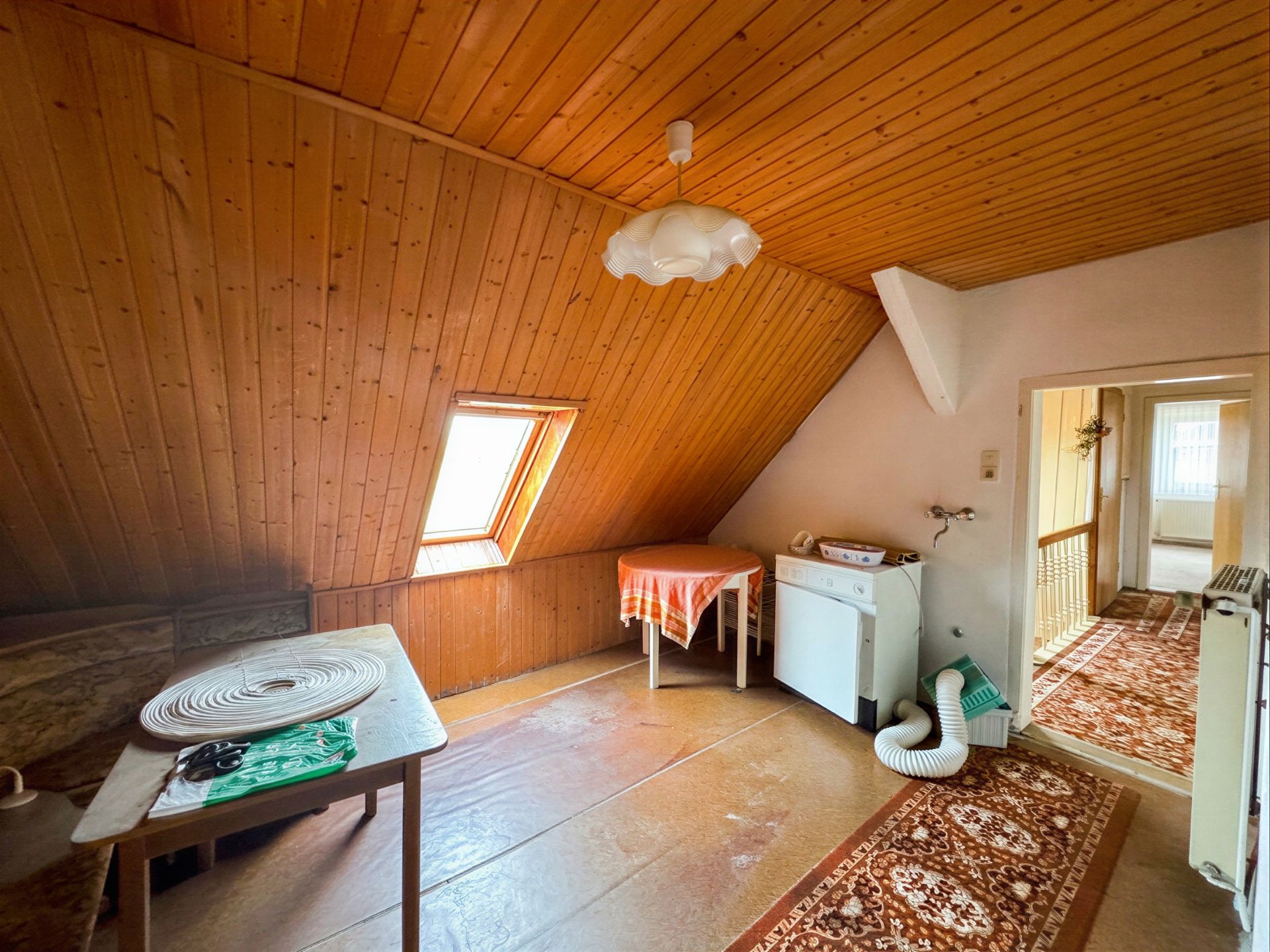 Dachzimmer mit Holzdecke, schrägem Fenster, Tisch, Waschmaschine und Teppichboden.