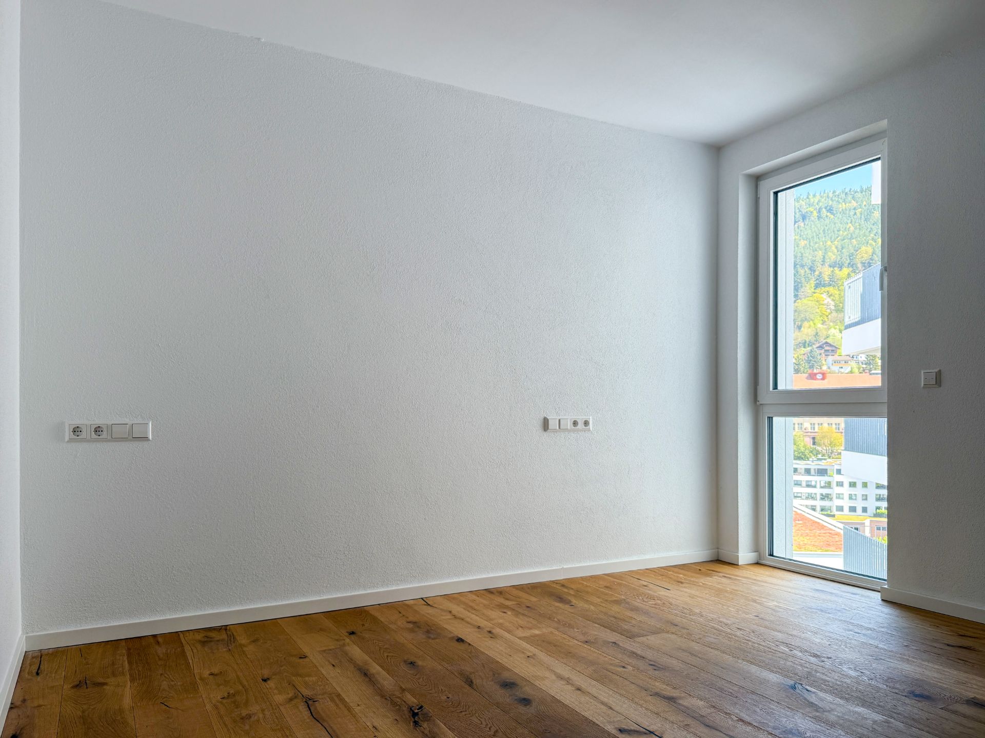 Leerer Raum mit Holzboden, weißer Wand und großem Fenster, das nach draußen zeigt.
