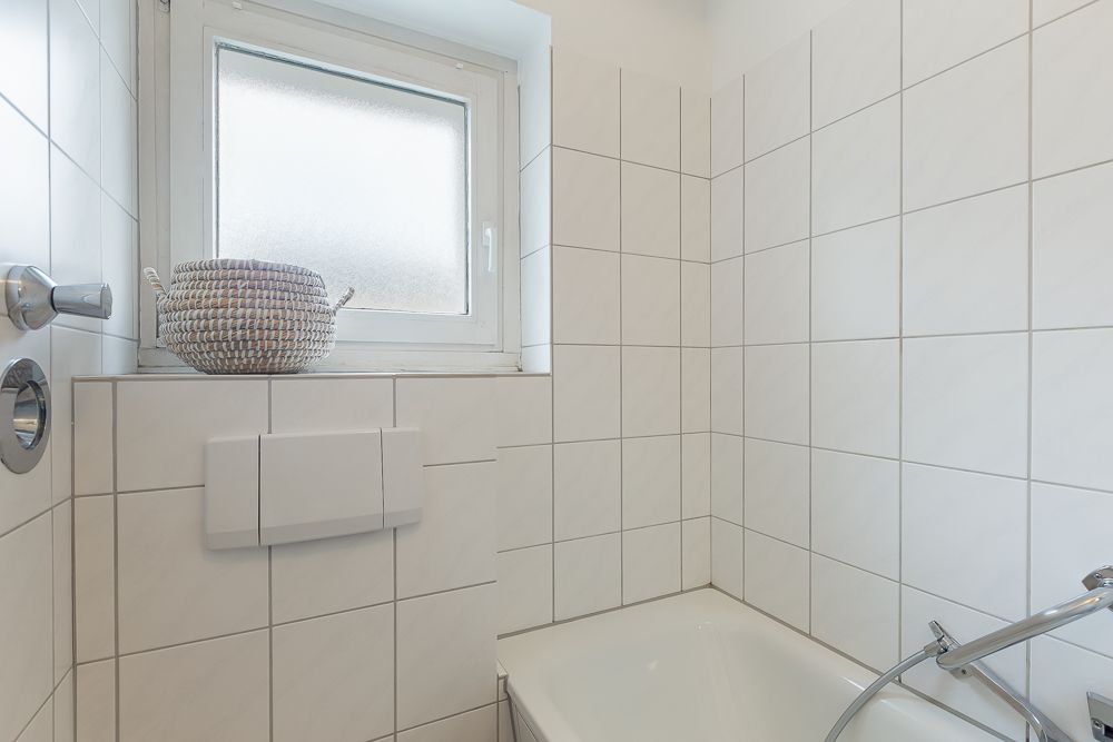 Helles Badezimmer mit weißen Fliesen, Fenster und Badewanne, Korb auf der Fensterbank.