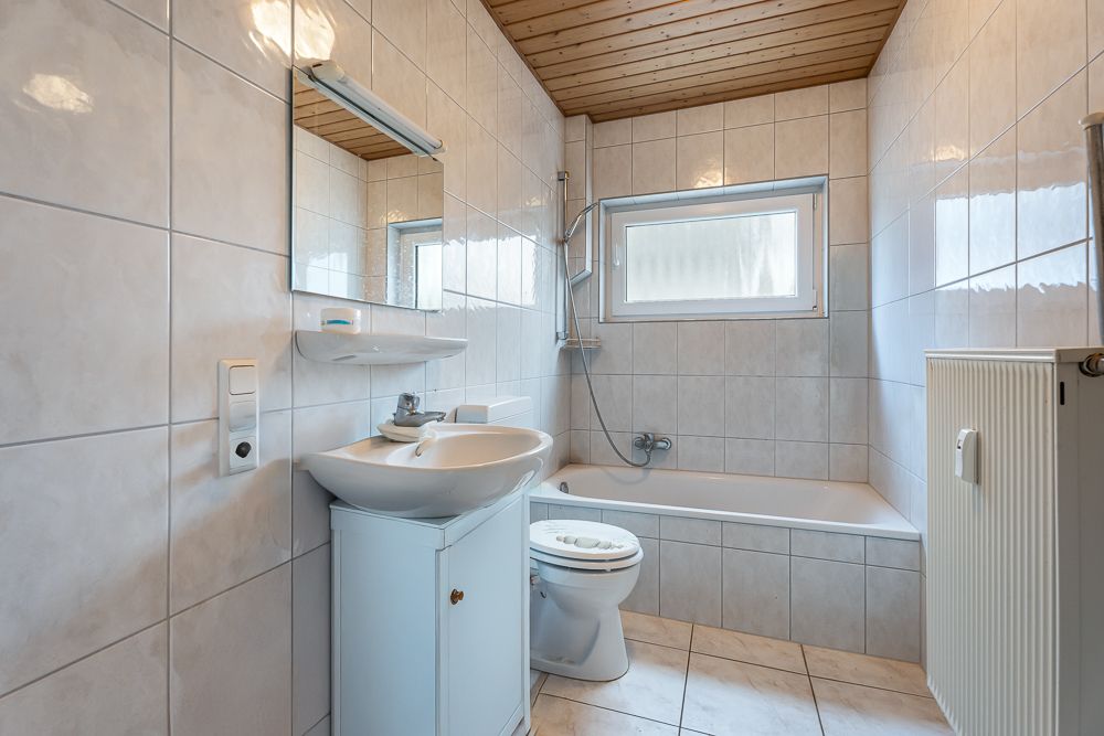 Helles Badezimmer mit Badewanne, Waschbecken, Toilette und Fenster, gefliest und Holzdecke.