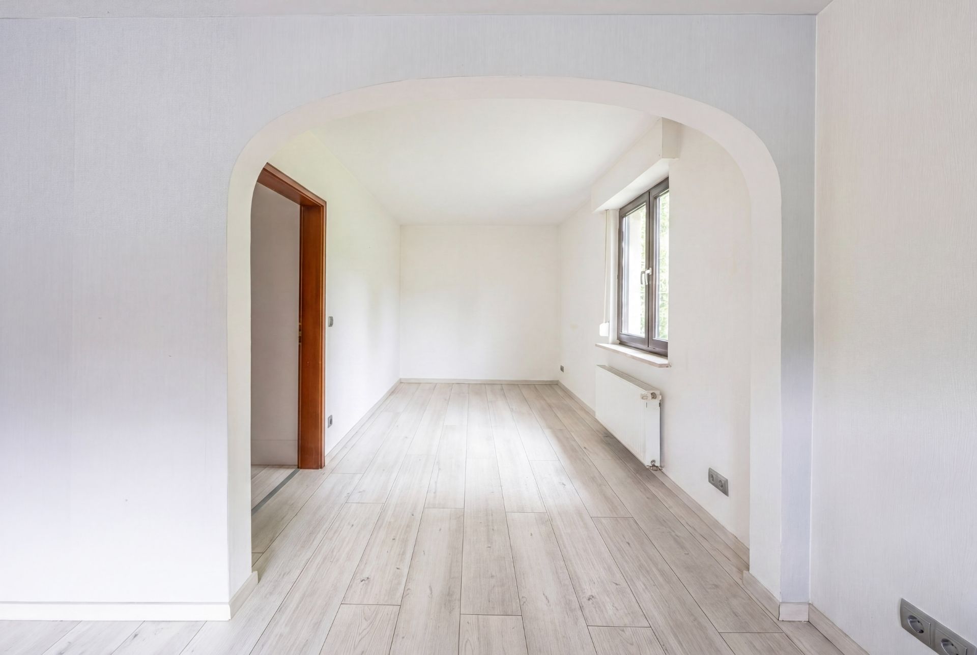 Helles, leeres Zimmer mit Holzboden, Bogenöffnung und großem Fenster auf der rechten Seite.