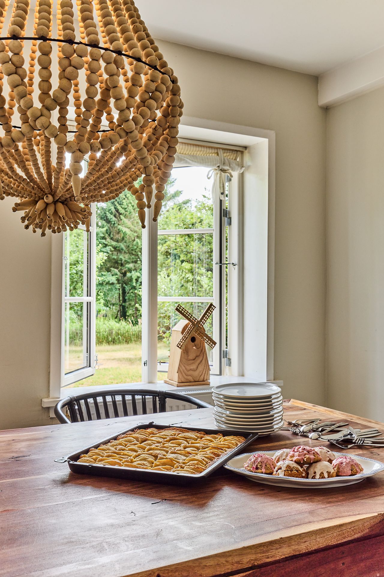 Großer Holztisch mit Gebäck und Tellern vor offenem Fenster und hölzernem Windmühlenmodell.