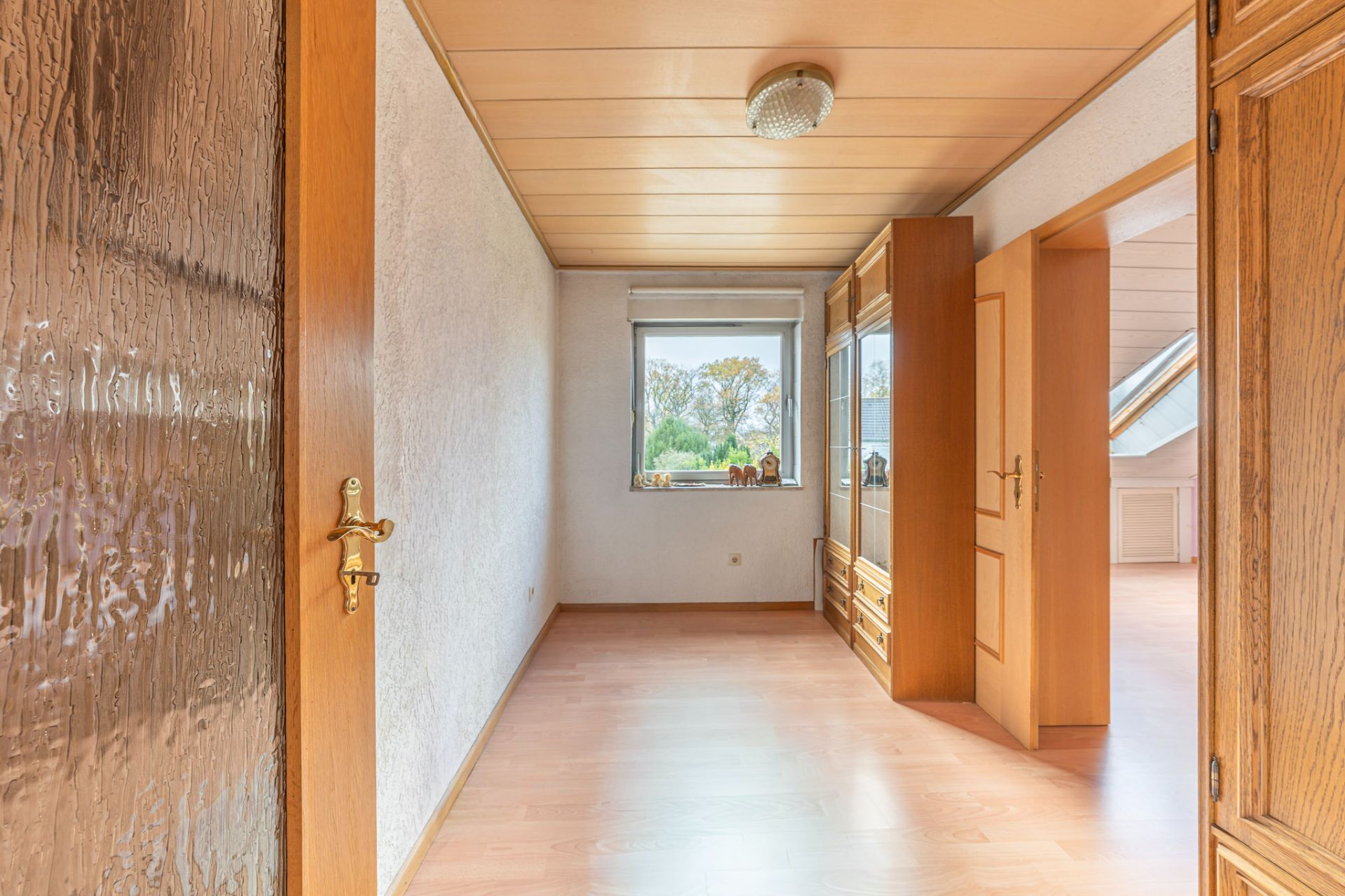 Heller Raum mit Holztür, Fenster und großem Holzschrank, Blick ins nächste Zimmer.
