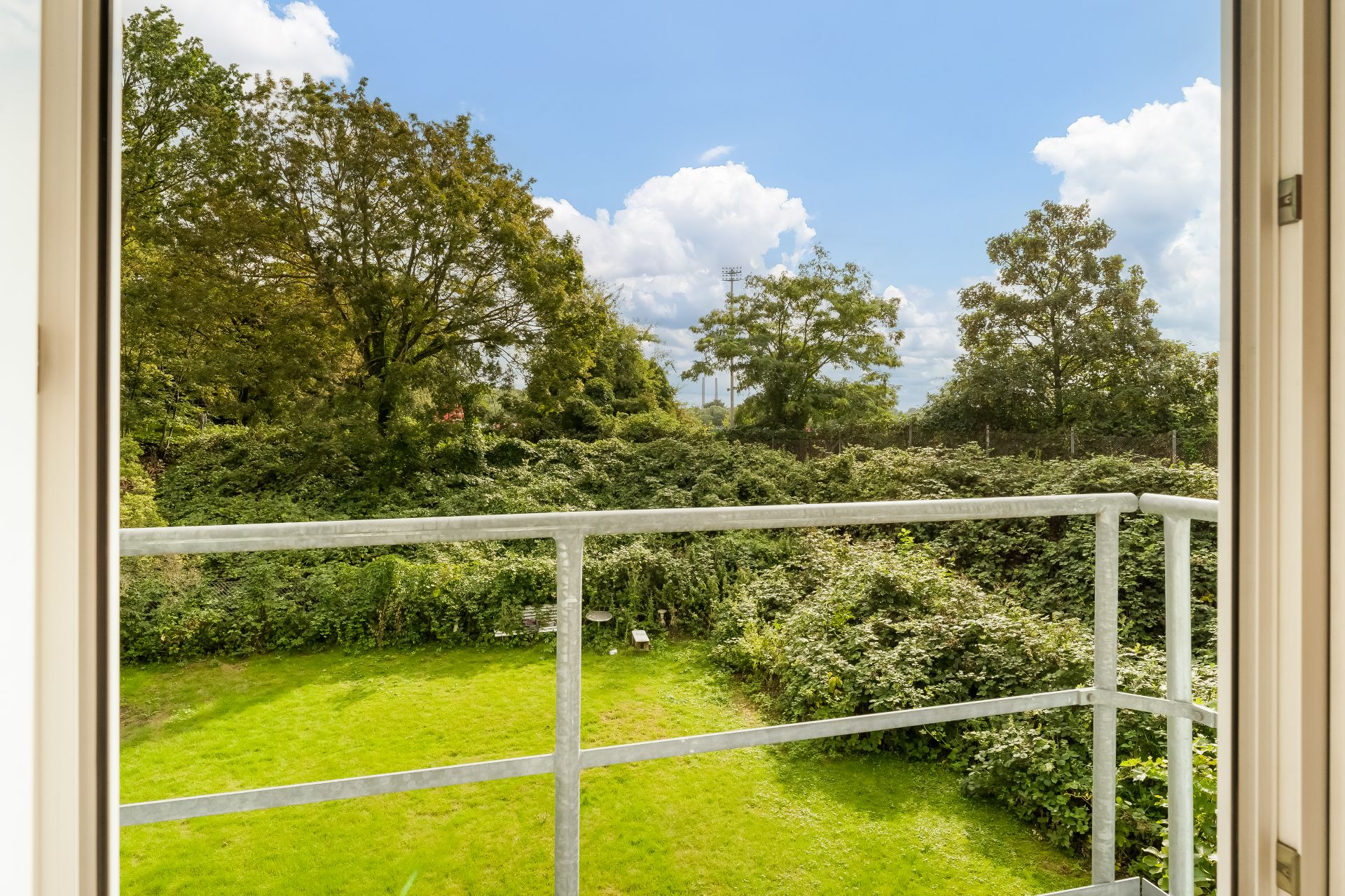 Blick vom Balkon auf einen grünen Garten mit Büschen und Bäumen bei Sonnenschein.
