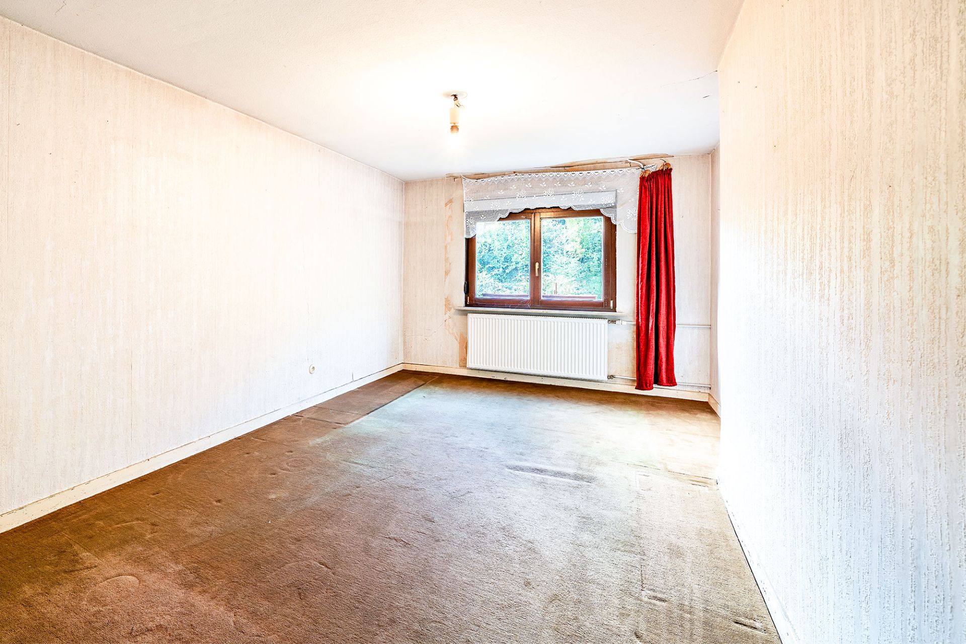 Leeres Zimmer mit Teppichboden, Fenster mit rotem Vorhang und Heizung darunter.