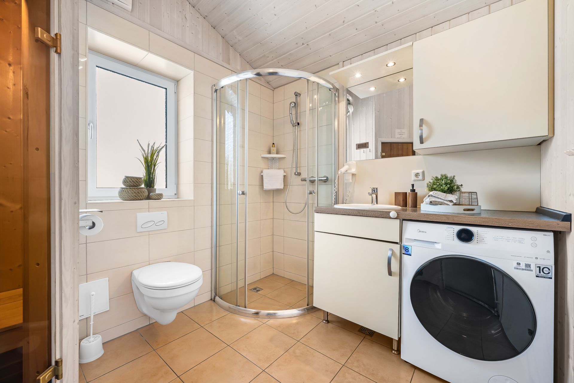 Modernes Badezimmer mit Eckdusche, Waschmaschine, Waschbecken und WC, helle Fliesen und Fenster.