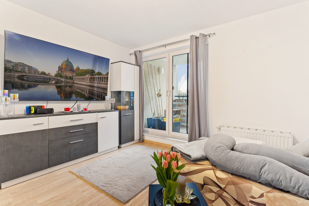 Modernes Wohnzimmer mit Sofa, Sideboard, Fernseher, Blumenstrauß und Zugang zu einem Balkon.