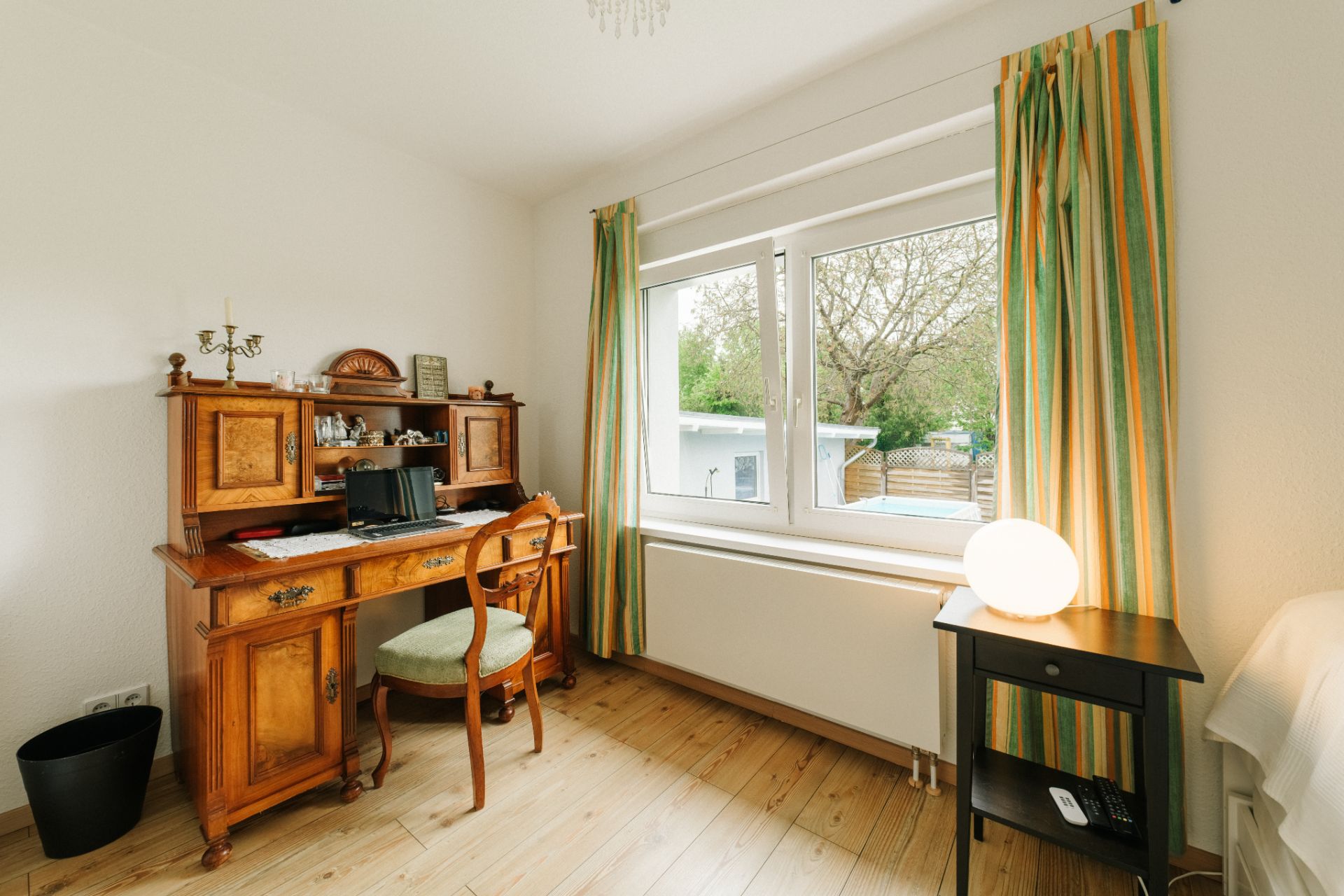 Helles Zimmer mit Holzschreibtisch, Stuhl, großem Fenster und Vorhängen, Lampe auf kleinem Tisch.