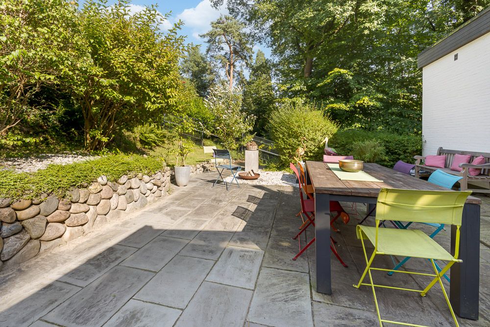 Gepflasterte Terrasse mit bunten Stühlen, Holztisch und viel Grün im Garten.
