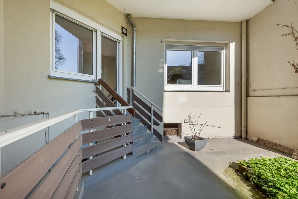 Kleiner, überdachter Außenbereich mit Treppe, Geländer, Fenstern und einem Blumentopf.