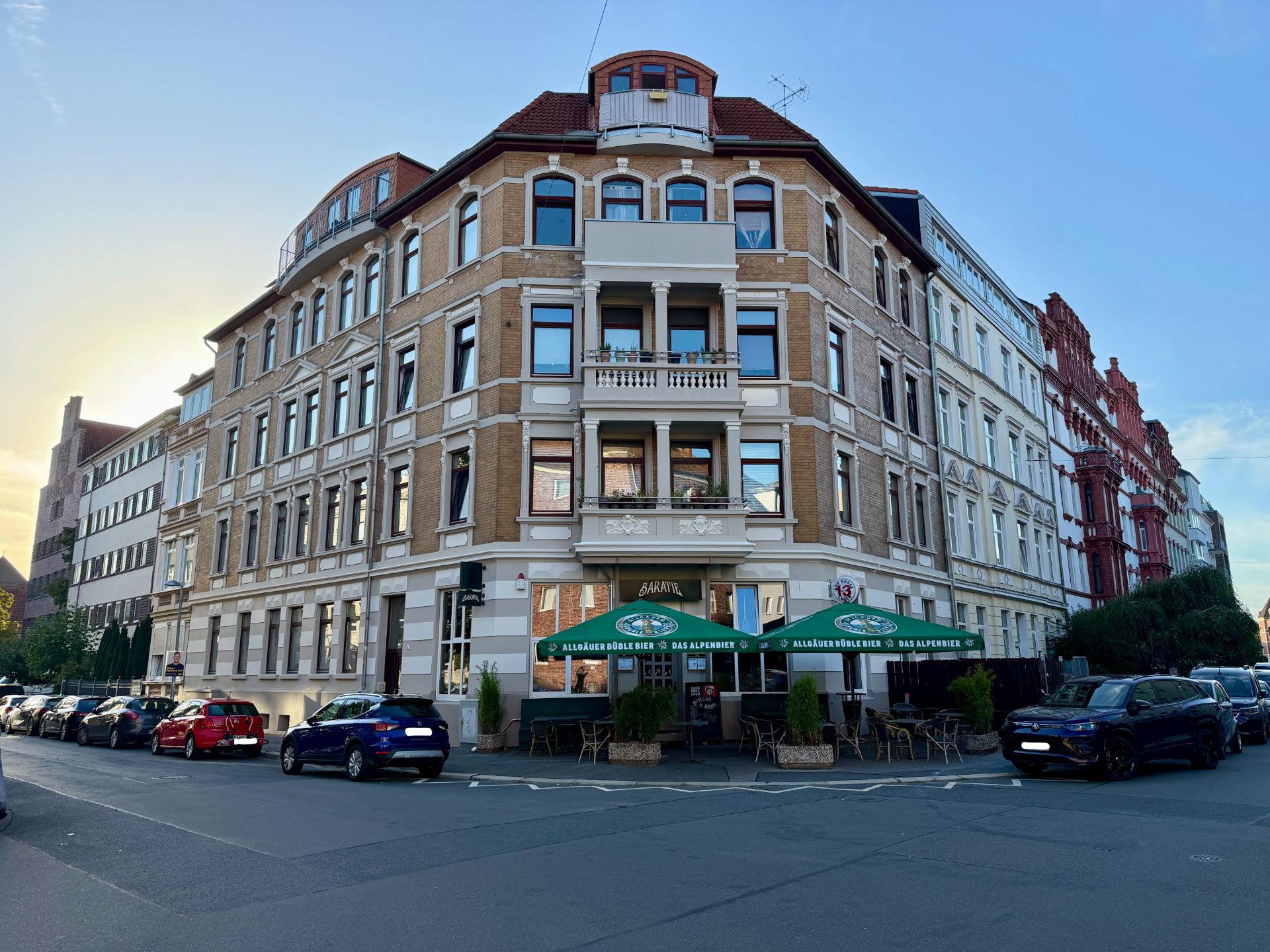 Eckhaus mit mehreren Etagen, Balkon und Café im Erdgeschoss, parkende Autos davor.