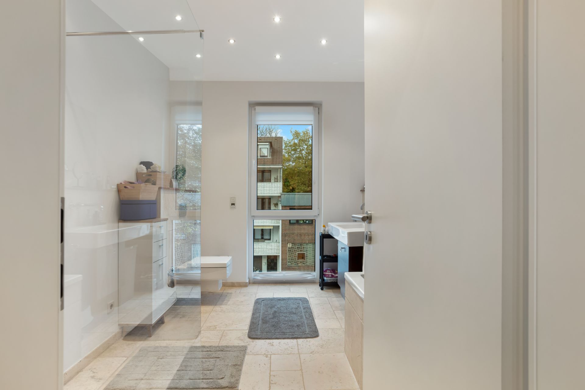 Modernes Badezimmer mit großem Fenster, beigen Fliesen und zwei Teppichen.