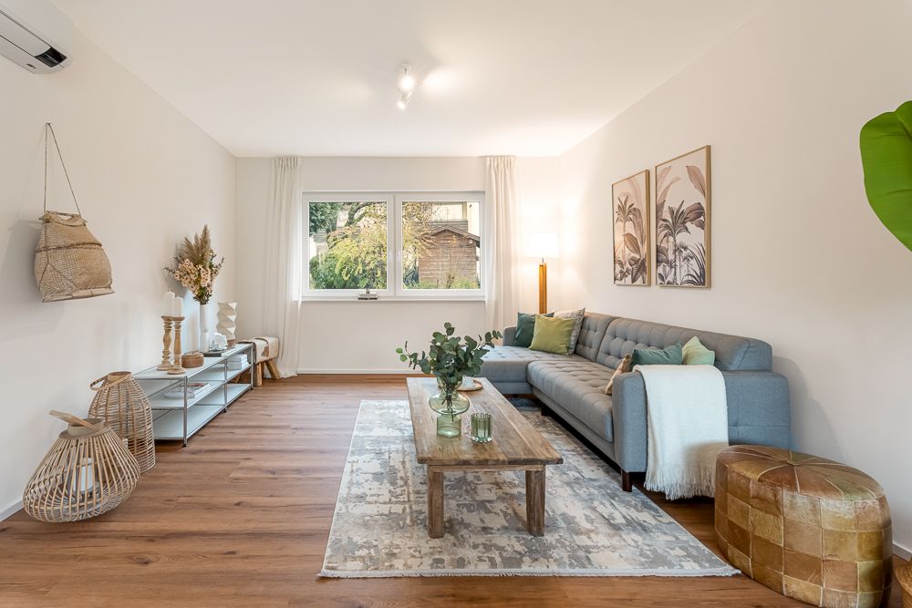Helles Wohnzimmer mit grauem Sofa, Holztisch, Teppich, Pflanzen und großem Fenster.