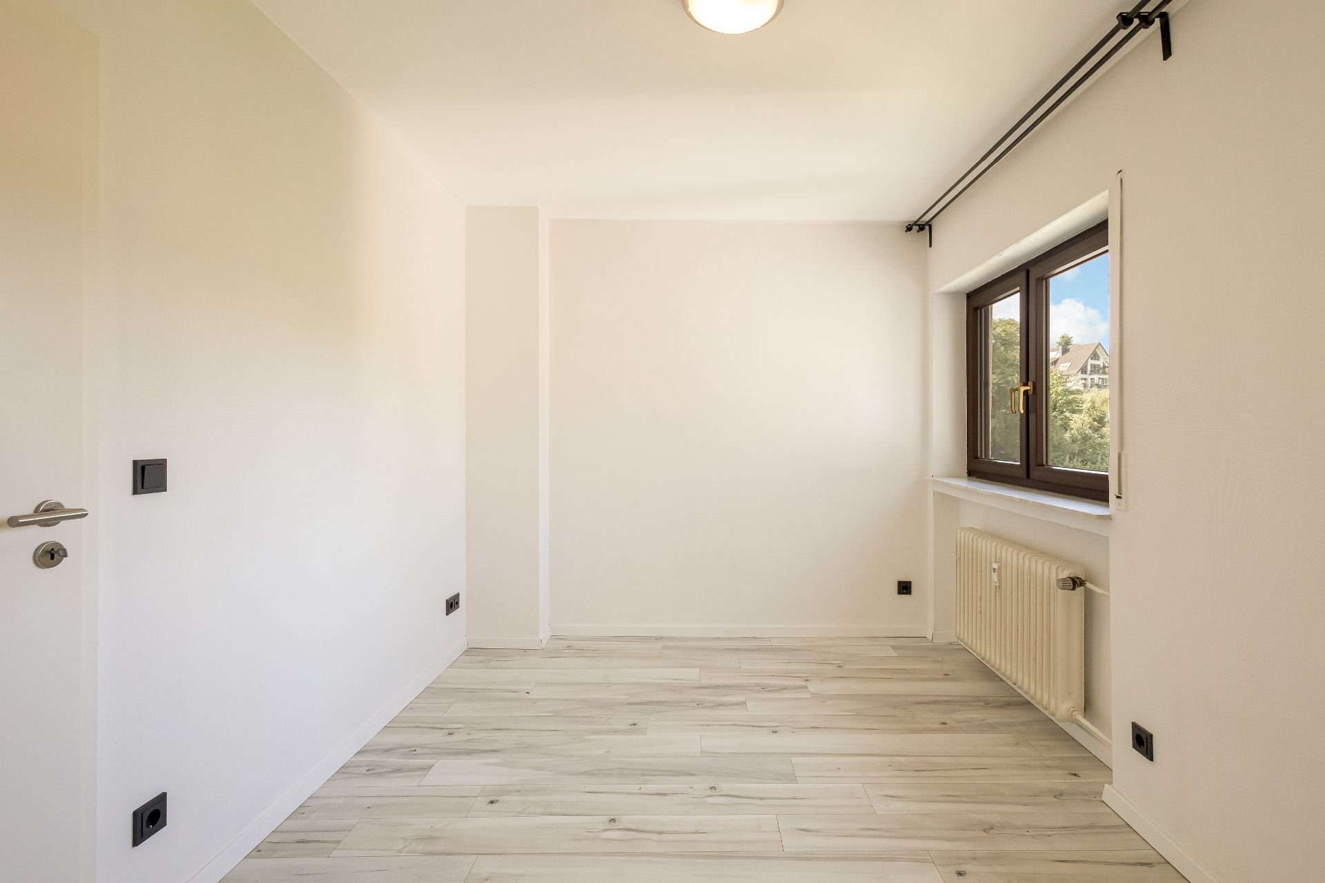 Helles, leeres Zimmer mit großem Fenster, Heizung und hellem Holzfußboden.