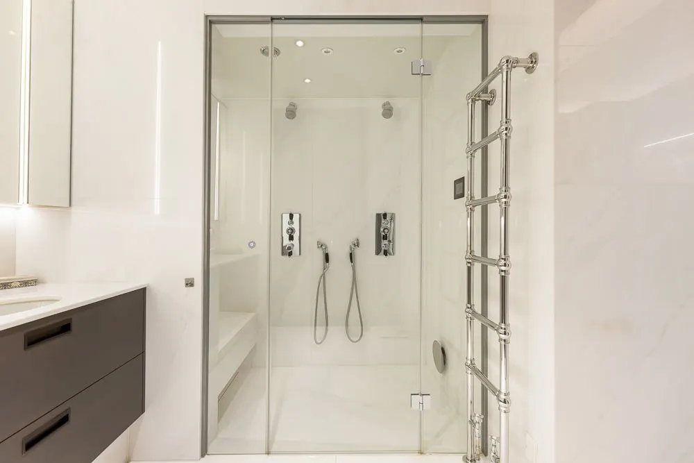 Modernes Badezimmer mit großer Glasdusche, zwei Duschköpfen und Handbrausen, Handtuchheizkörper an der Wand.